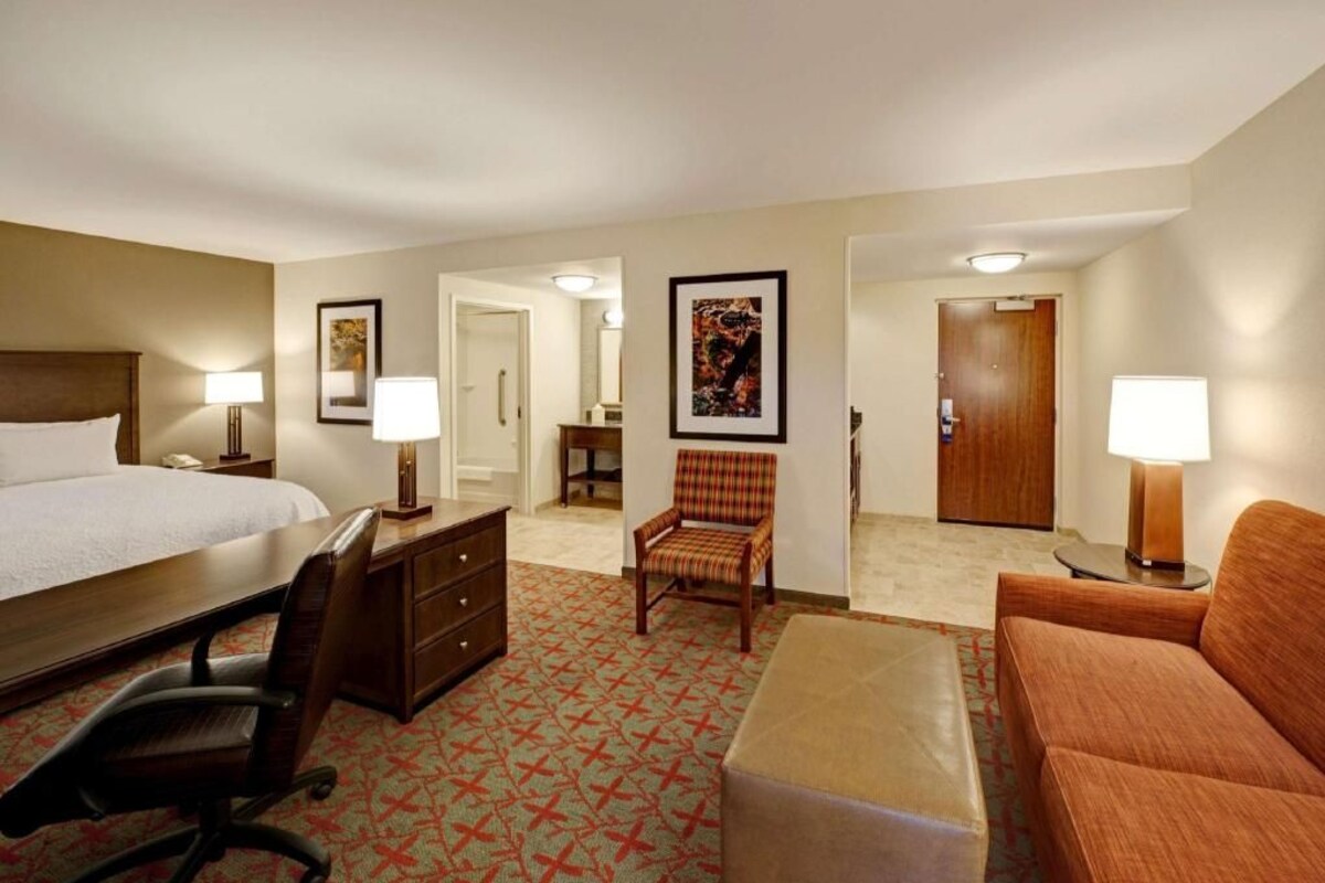 Habitación de hotel con cama, escritorio, silla, sofá y una mesita. Hay dos lámparas, una puerta y un cuadro en la pared.