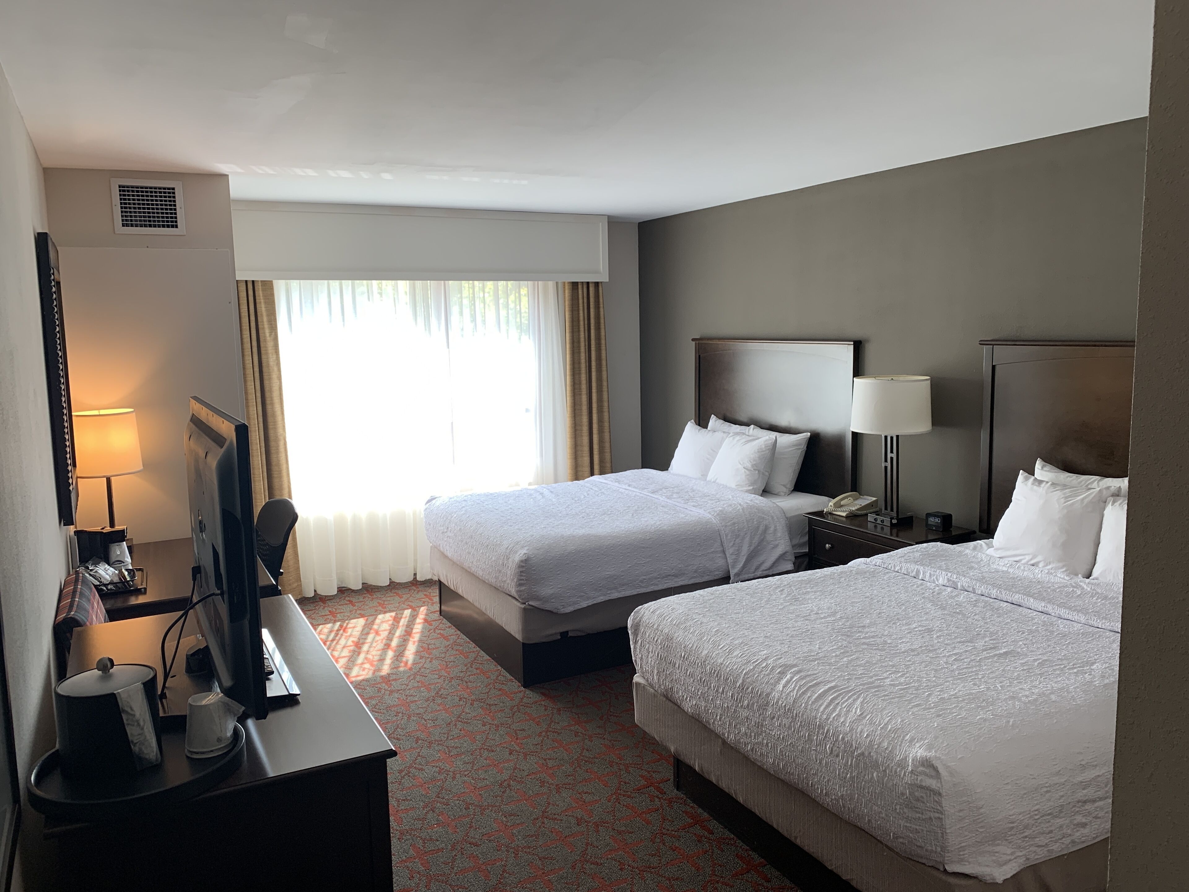 A hotel room with two beds, a desk with a computer and a lamp, a bedside table with a phone and a lamp, and a window with curtains.