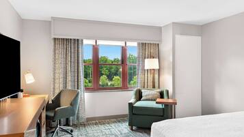 Une chambre d’hôtel avec un lit, un bureau doté d’une lampe, une chaise, une fenêtre avec des rideaux et une vue sur les arbres.