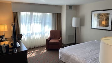 A hotel room with a bed, a chair, a desk, a lamp, and a window with curtains.