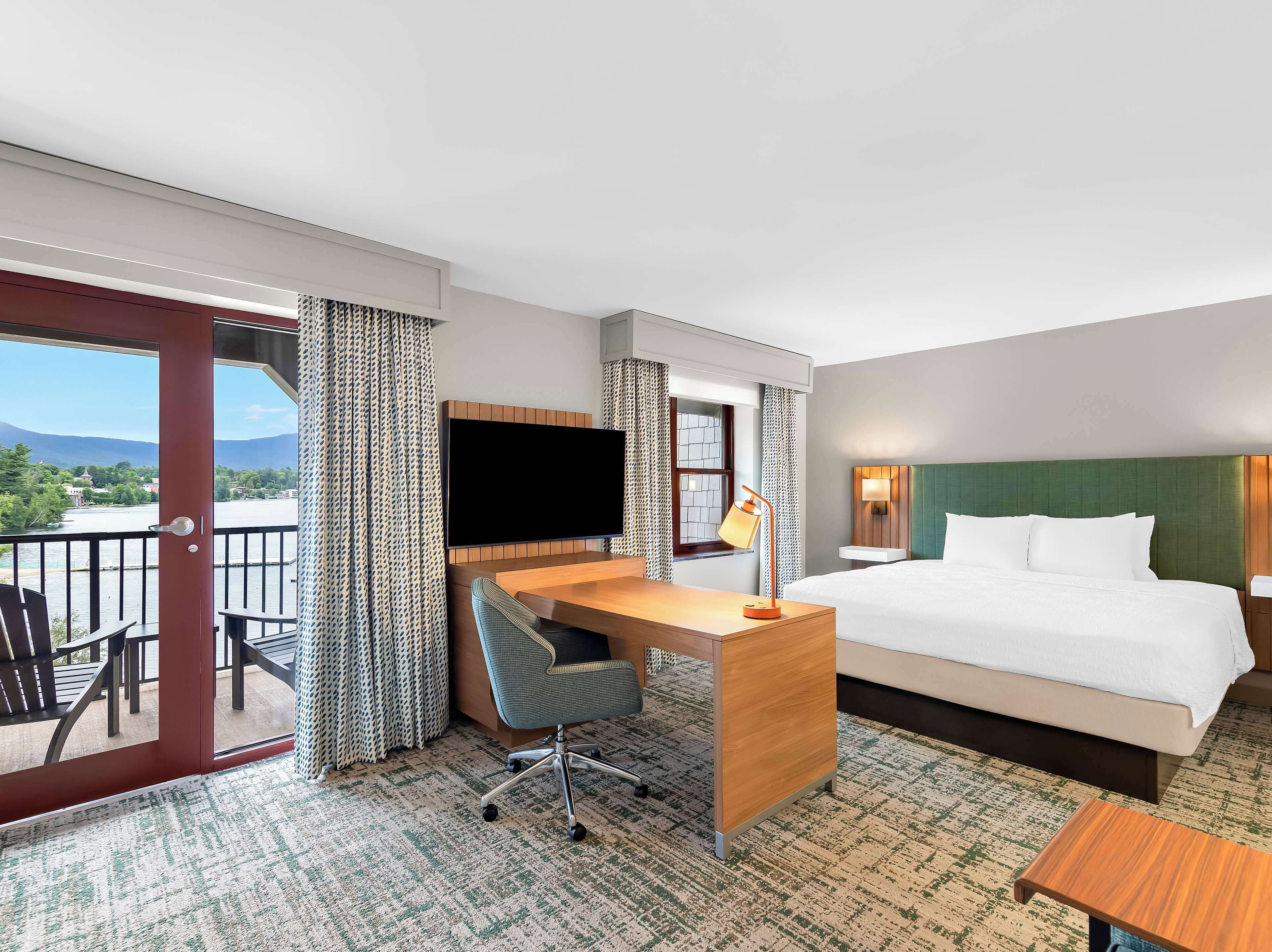 Een hotelkamer met een bed, bureau, stoel, televisie en uitzicht op een meer en bergen.