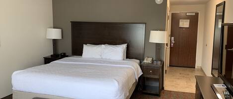 A hotel room with a large bed, two bedside tables with lamps, a wooden headboard, a small desk with a mirror, and a door leading to another room.