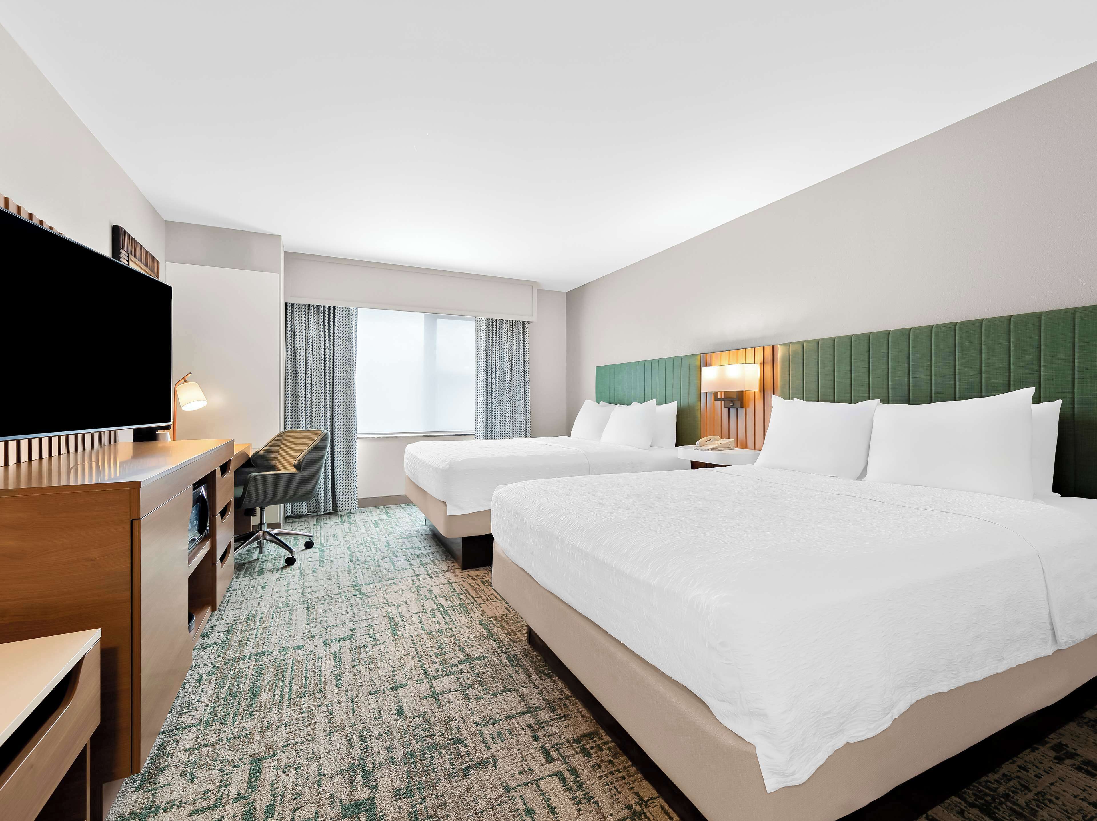 A hotel room with two beds, a desk with a flat-screen TV, a chair, a lamp, and a window with curtains.