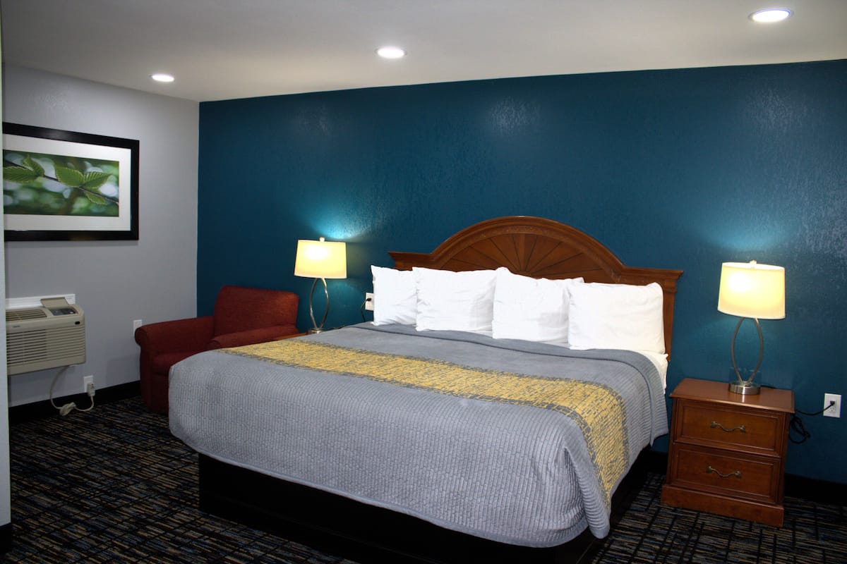 A hotel room with a bed, two bedside lamps, a red chair, a wooden nightstand, and a wall-mounted air conditioner.