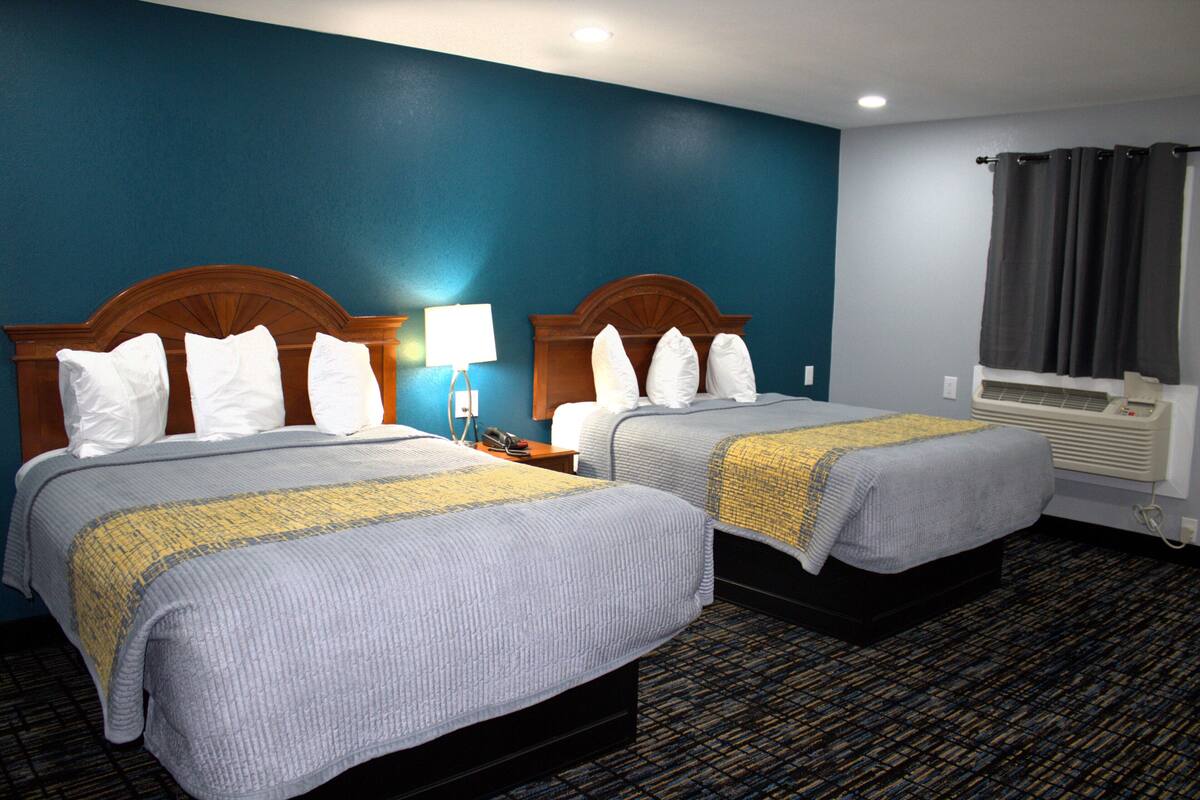 A hotel room with two beds, a nightstand with a lamp, a window with curtains, and an air conditioning unit.