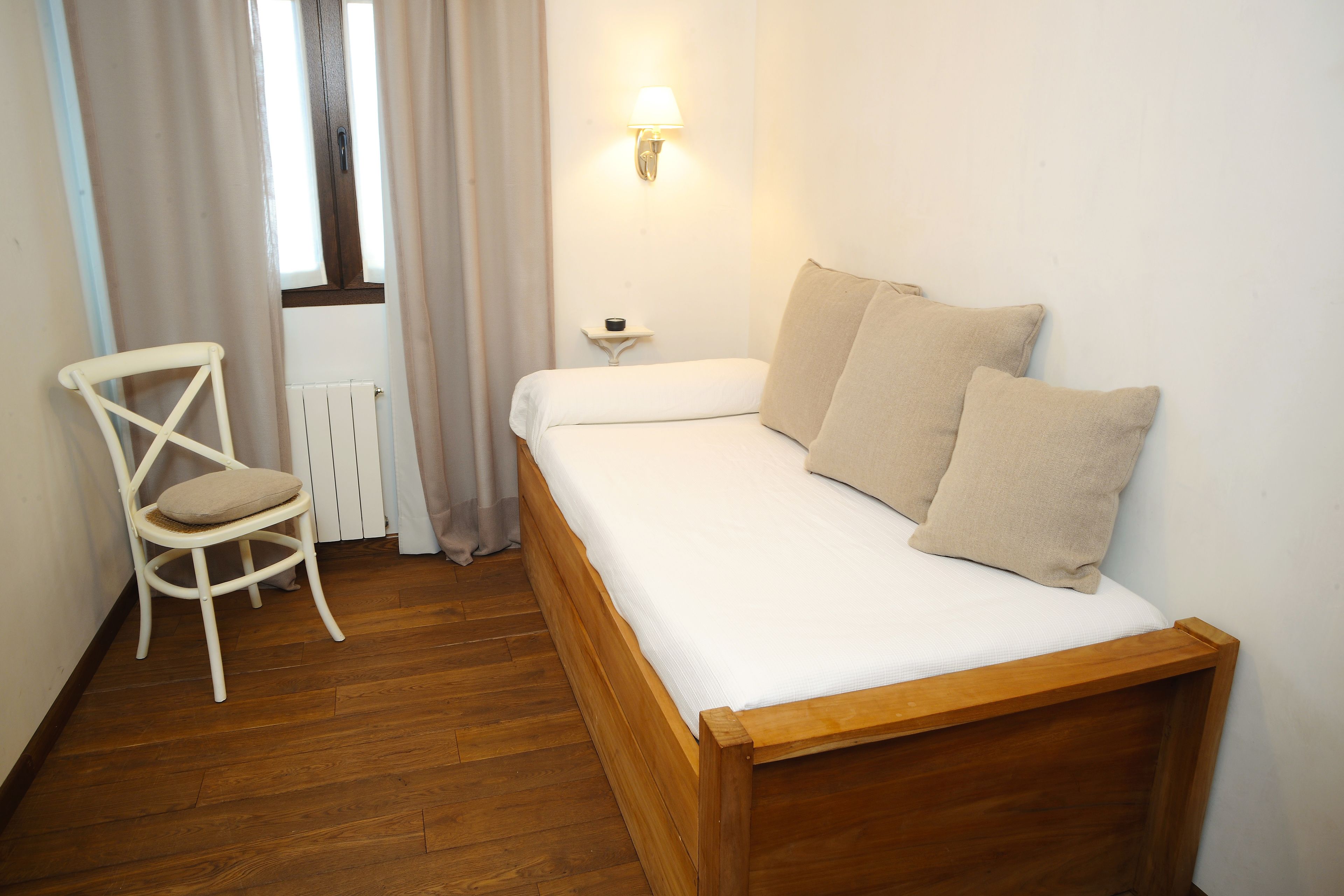 Un lit simple avec un cadre en bois et des oreillers de couleur beige, une chaise blanche avec un coussin, une table de chevet avec une lampe, et une fenêtre avec des rideaux.