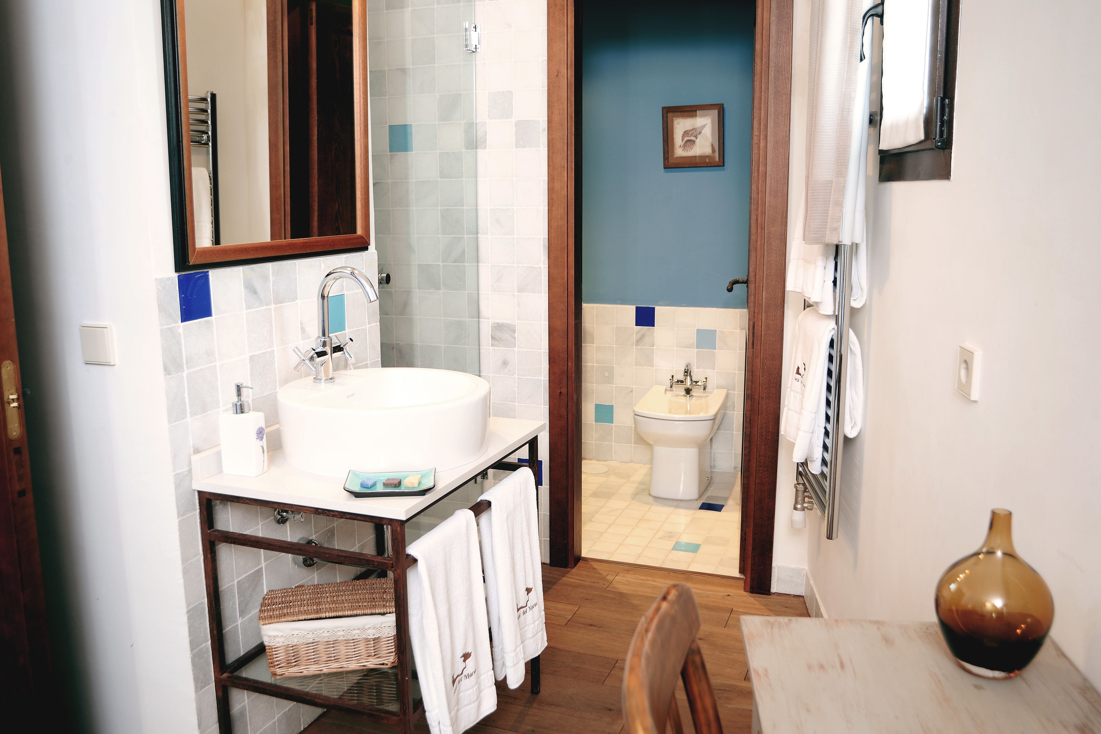 Un baño con lavabo, espejo e inodoro. Hay una silla de madera y un florero sobre una mesa.
