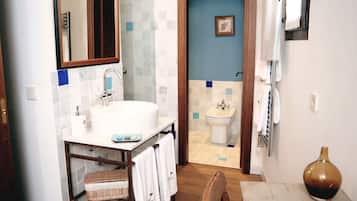 Un baño con lavabo, espejo e inodoro. Hay una silla de madera y un florero sobre una mesa.