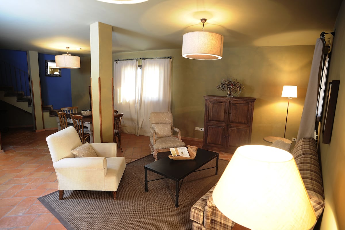 A living room with a sofa, armchair, coffee table, and a lamp. There is a staircase leading to another room, a cabinet, and a window with curtains.