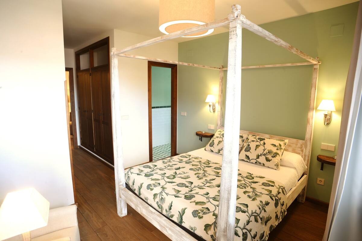 A four-poster bed with floral bedding, two bedside lamps, and a wooden floor.