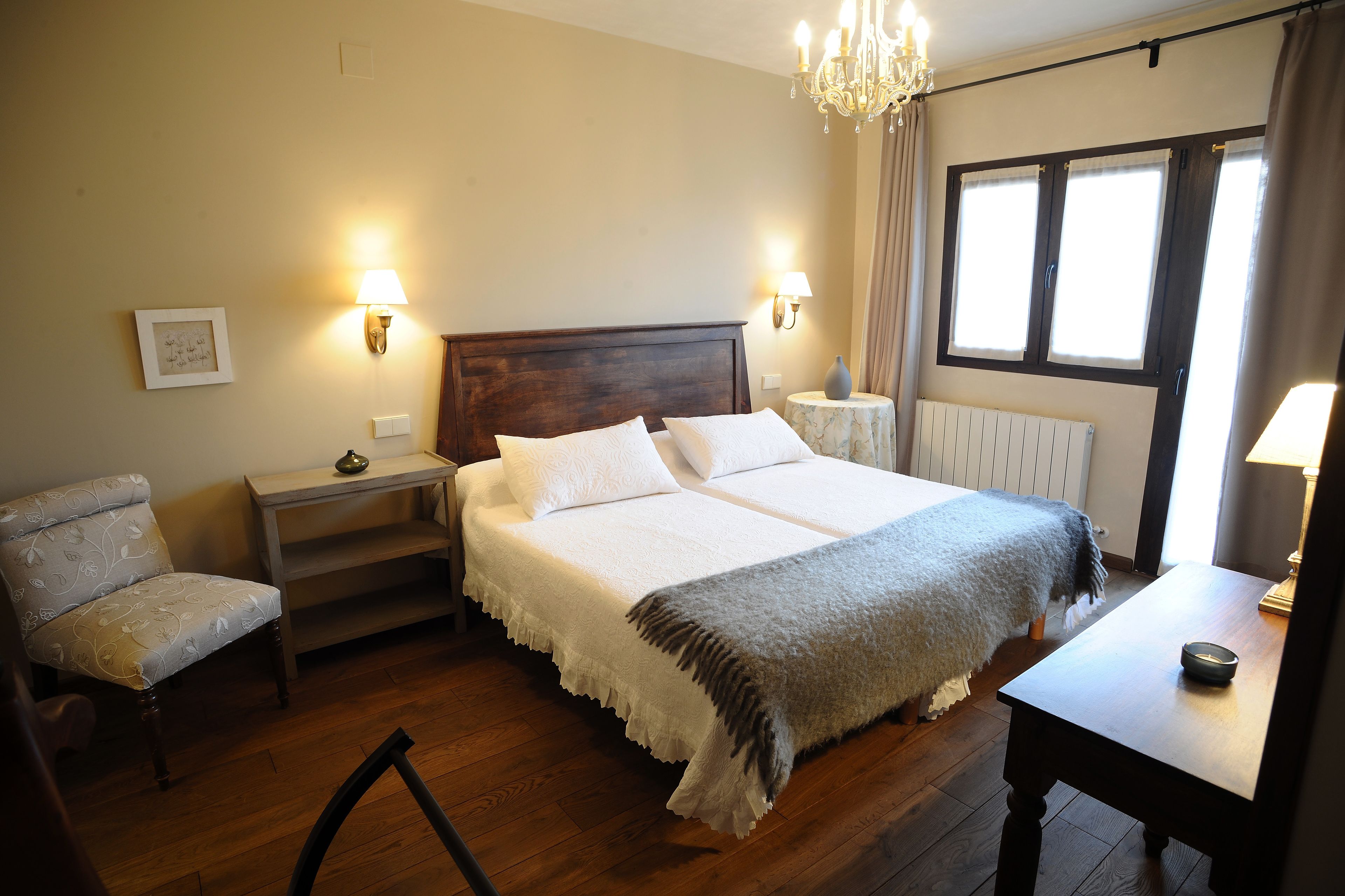 Un dormitorio con cama doble, lámparas de noche, una silla, una mesa y un ventanal con cortinas.