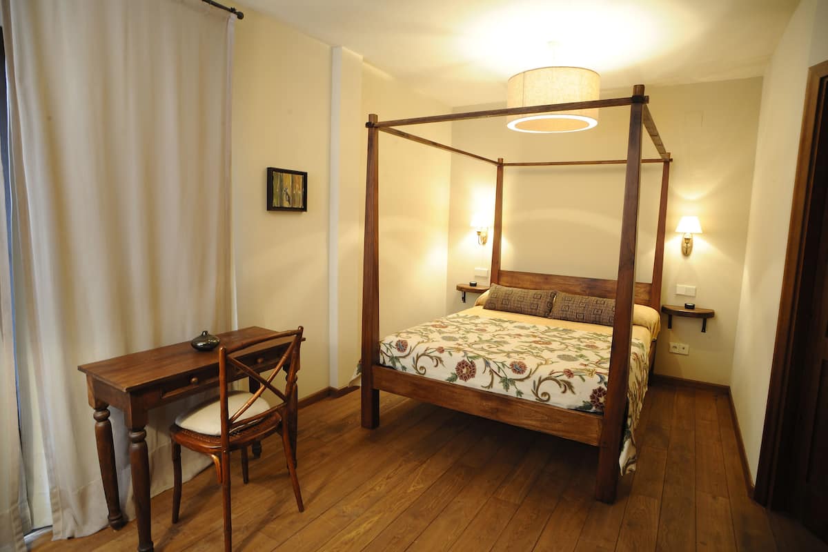A four-poster bed with a floral bedspread, a wooden desk with a chair, and two wall-mounted lamps.