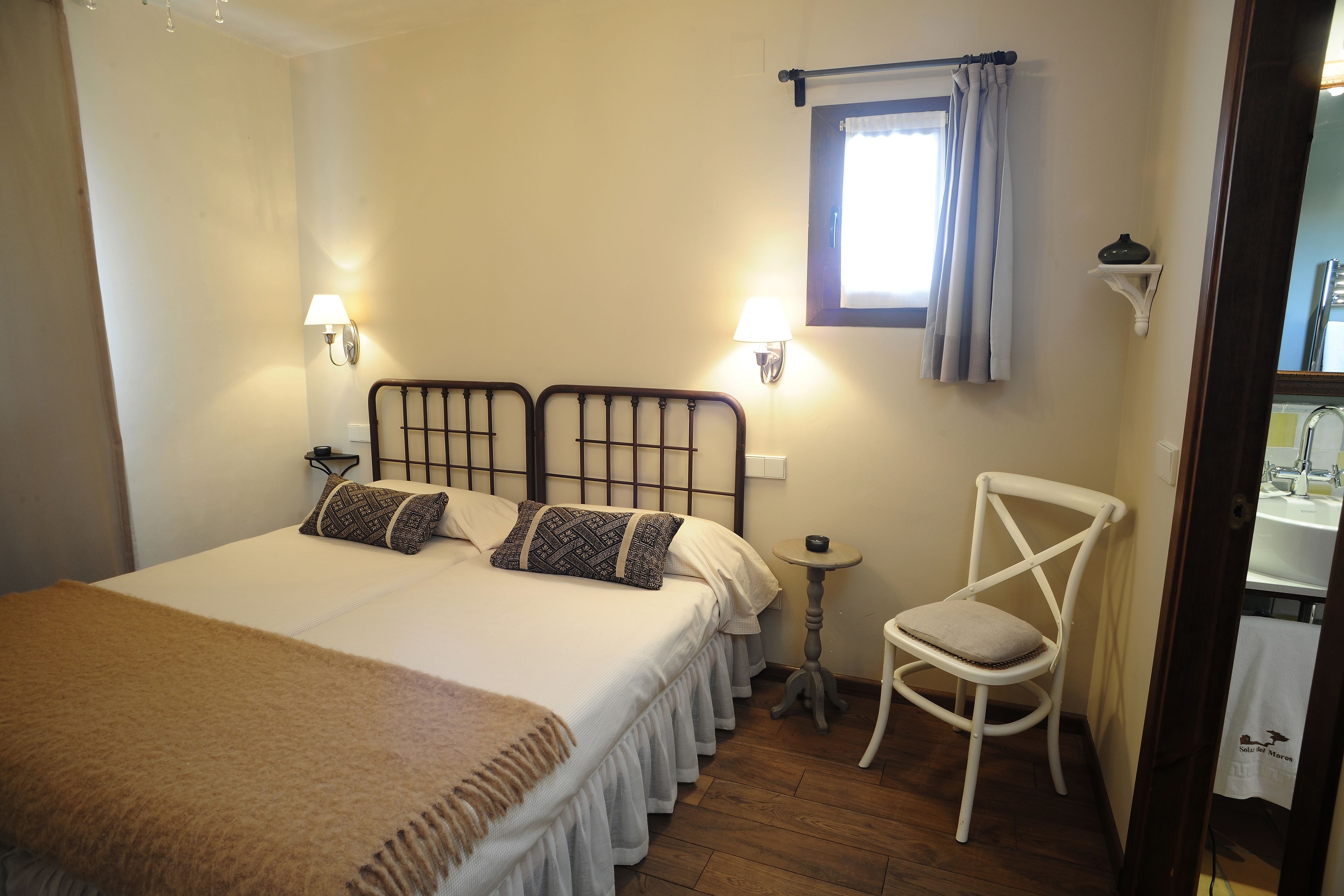 A bedroom with a large bed, two bedside lamps, a small table, a chair, and a window with curtains.