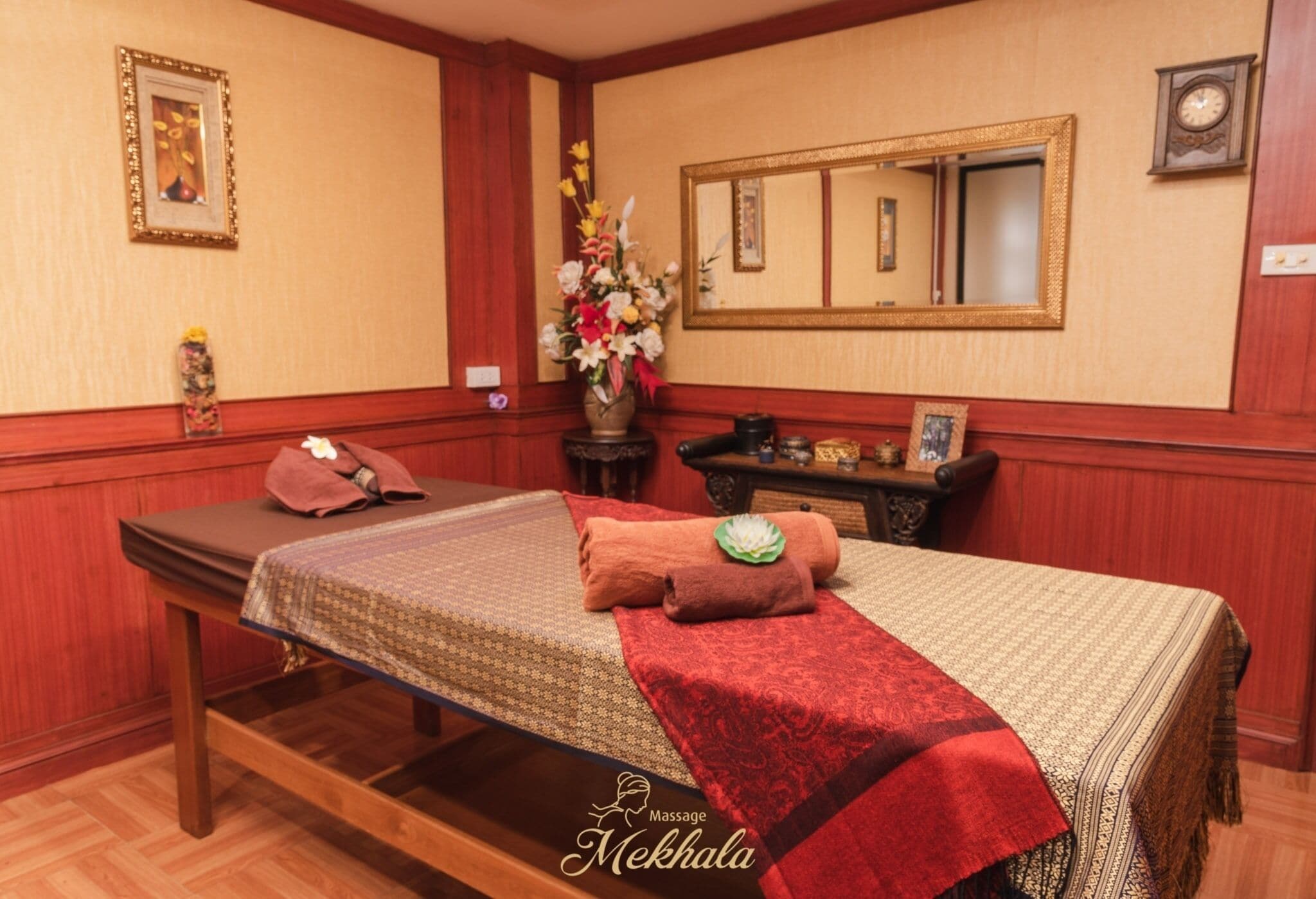 A massage room with a bed, a massage table, a mirror, and a vase with flowers.