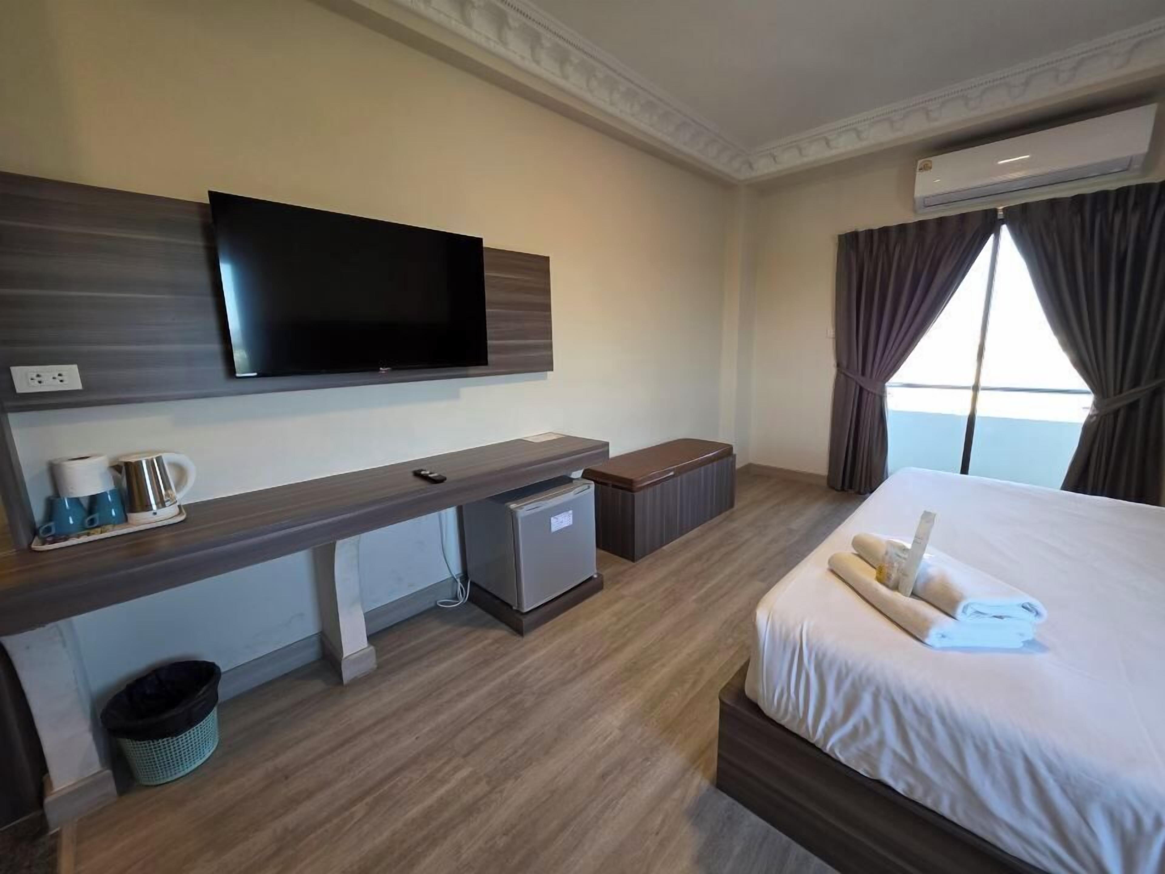 Habitación de hotel con cama, televisor montado en la pared, un escritorio pequeño con computadora, un cesto de basura y una ventana con cortinas.
