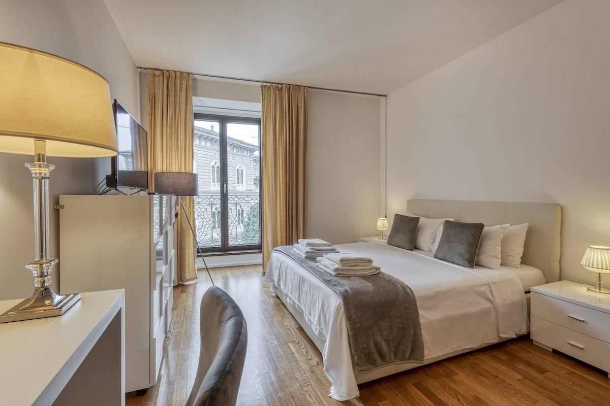 Habitación de hotel con cama, mesitas de noche, escritorio, silla, lámpara y vista a un edificio a través de una ventana.