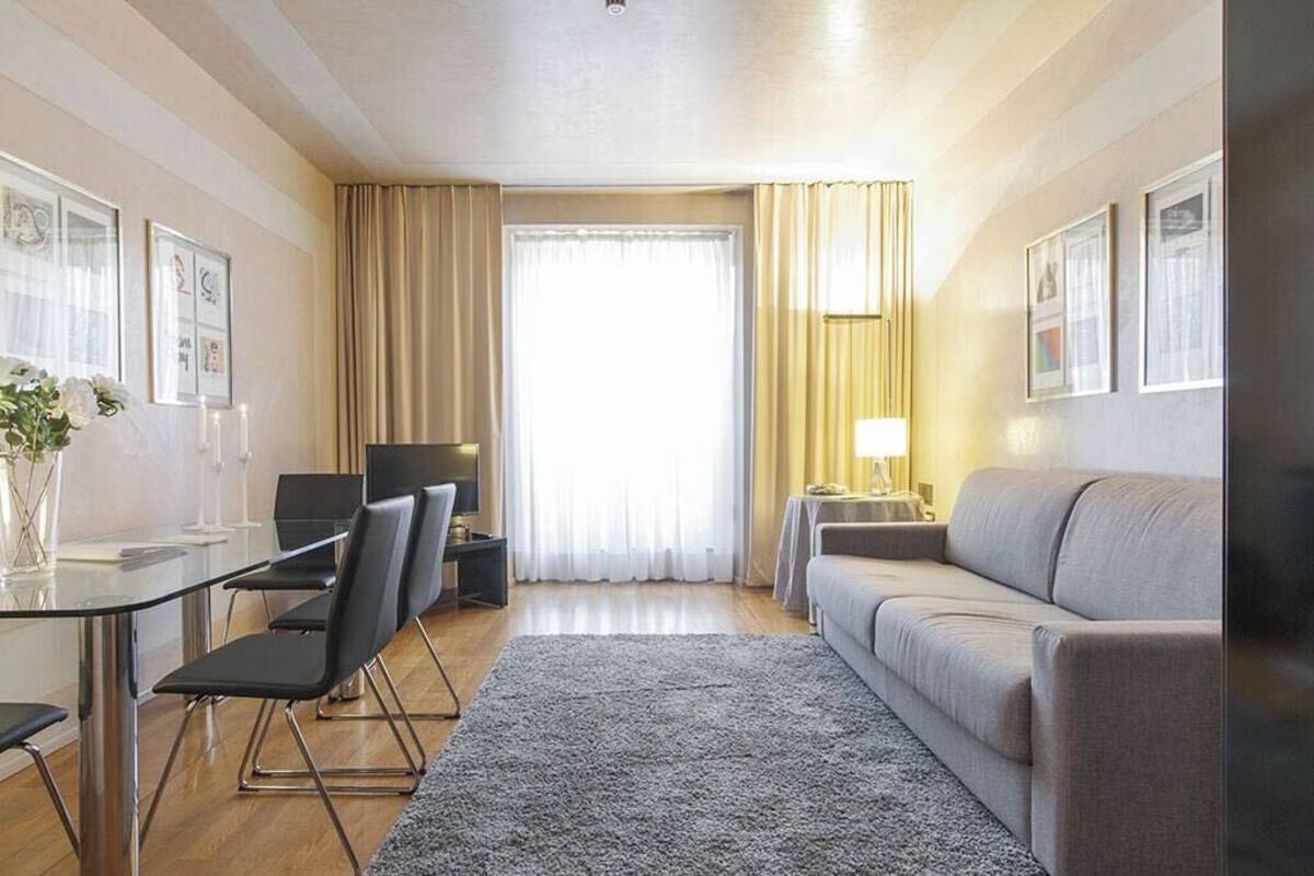 Habitación de hotel moderna con una mesa de comedor de cristal, sillas negras, un sofá gris, una mesita auxiliar con lámpara y un ventanal con cortinas.