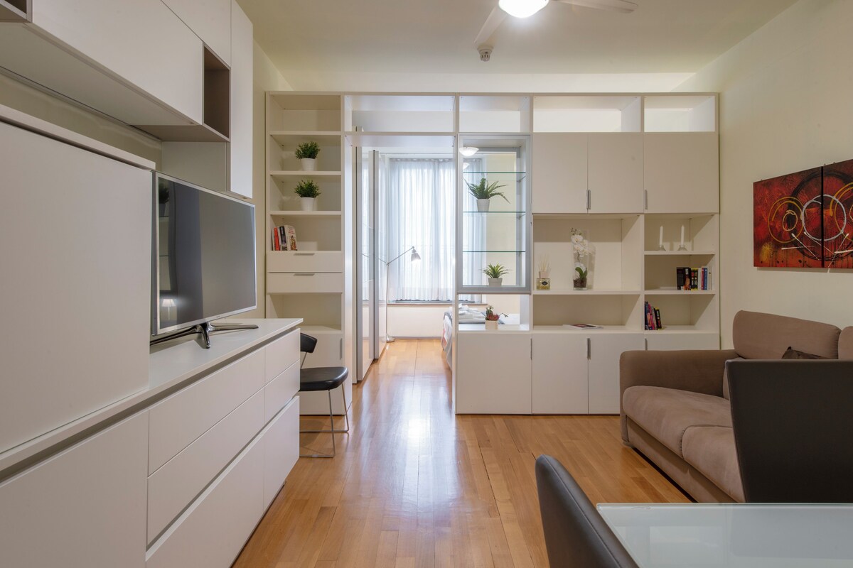 Een moderne woonkamer met een flatscreen televisie, een bank, een eettafel en een keuken met kastjes en een raam.