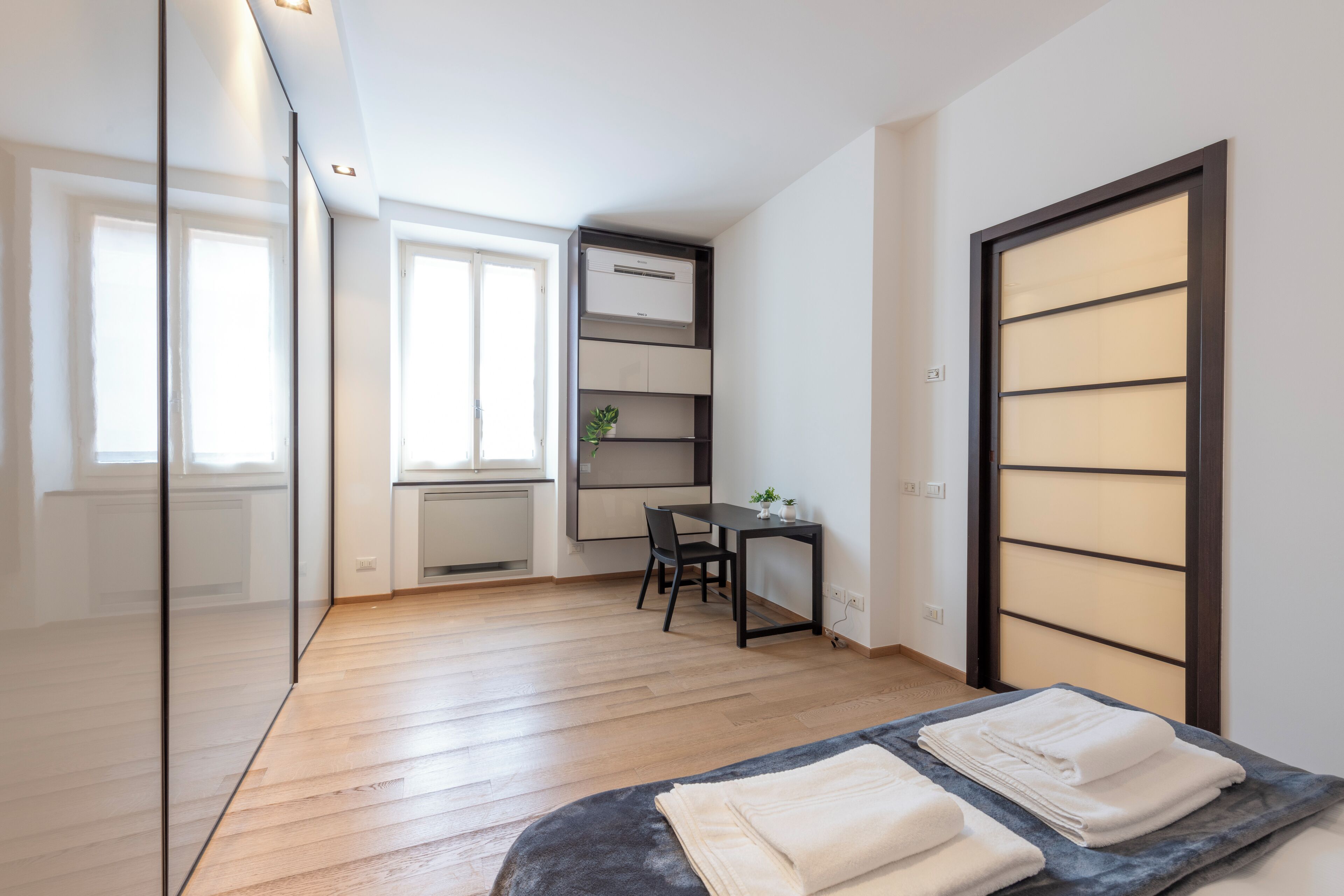 A modern bedroom with a wooden floor, a bed with neatly folded white towels, a desk with a chair, and a large window.