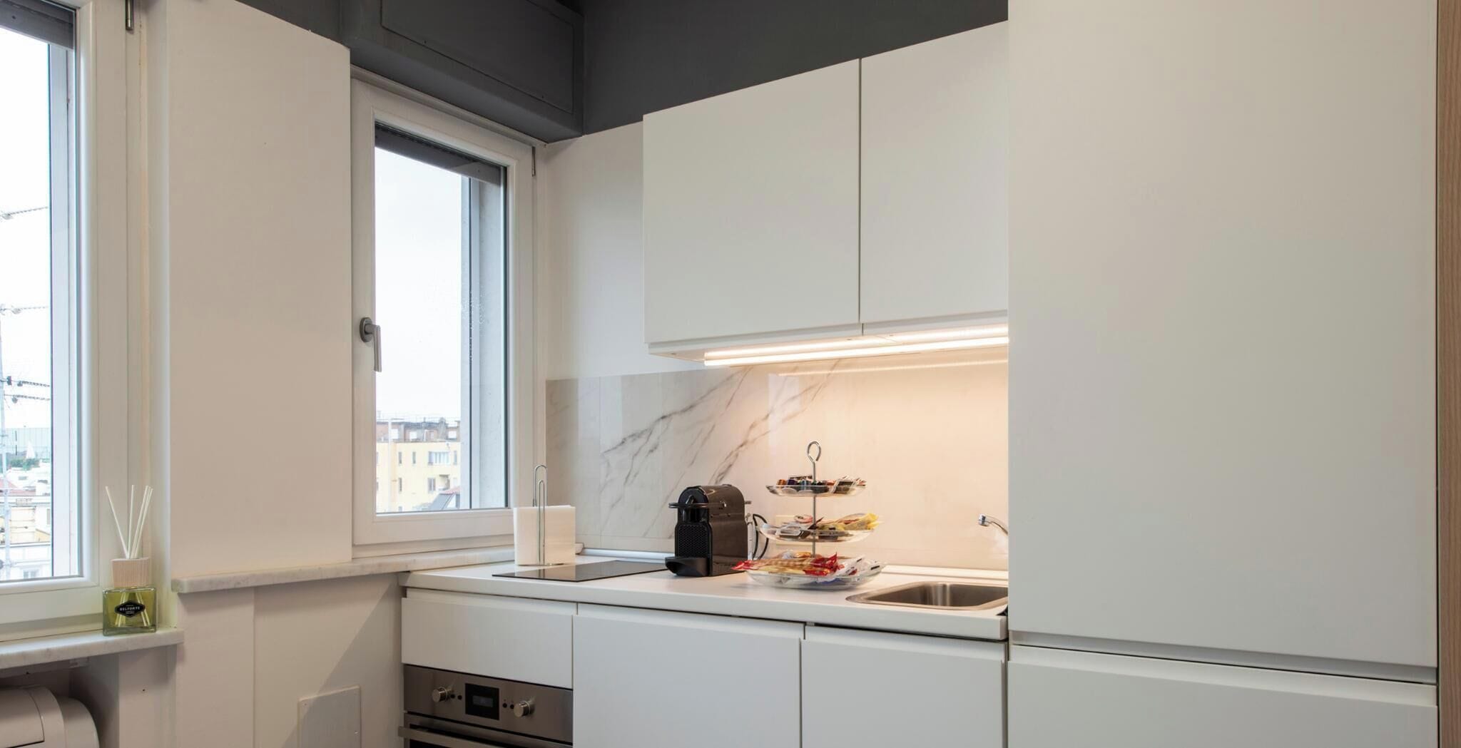 A modern kitchen with white cabinetry, a built-in oven, a coffee machine, and a window providing a view of the city.