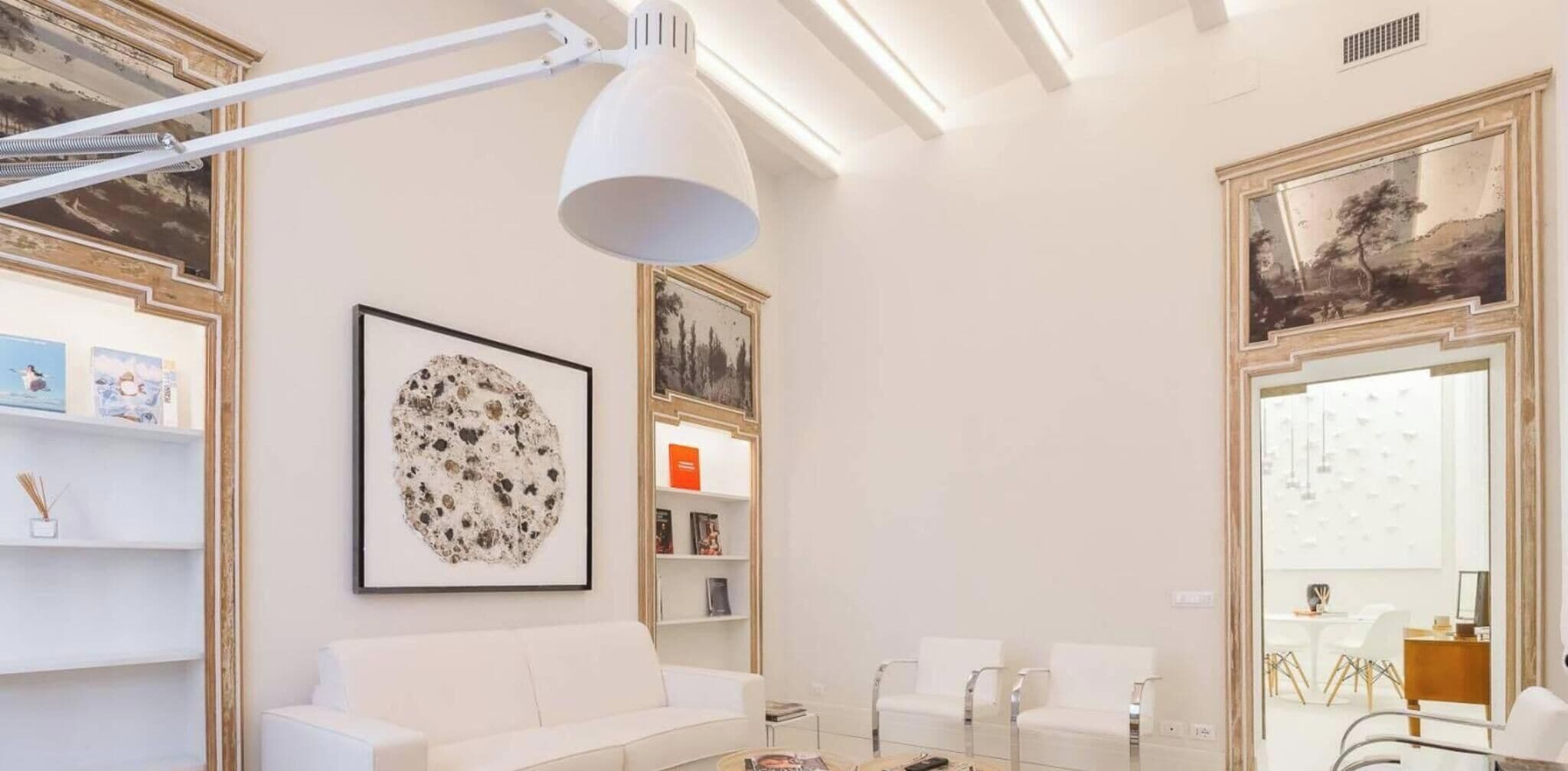 A modern living room with a white sofa, a coffee table, and a large abstract painting. There are bookshelves with books and decorative items, and a large mirror reflecting the room.