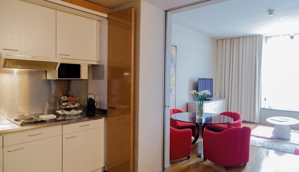 A modern kitchen with white cabinets and a microwave, a dining area with a glass table and red chairs, and a large window with curtains.