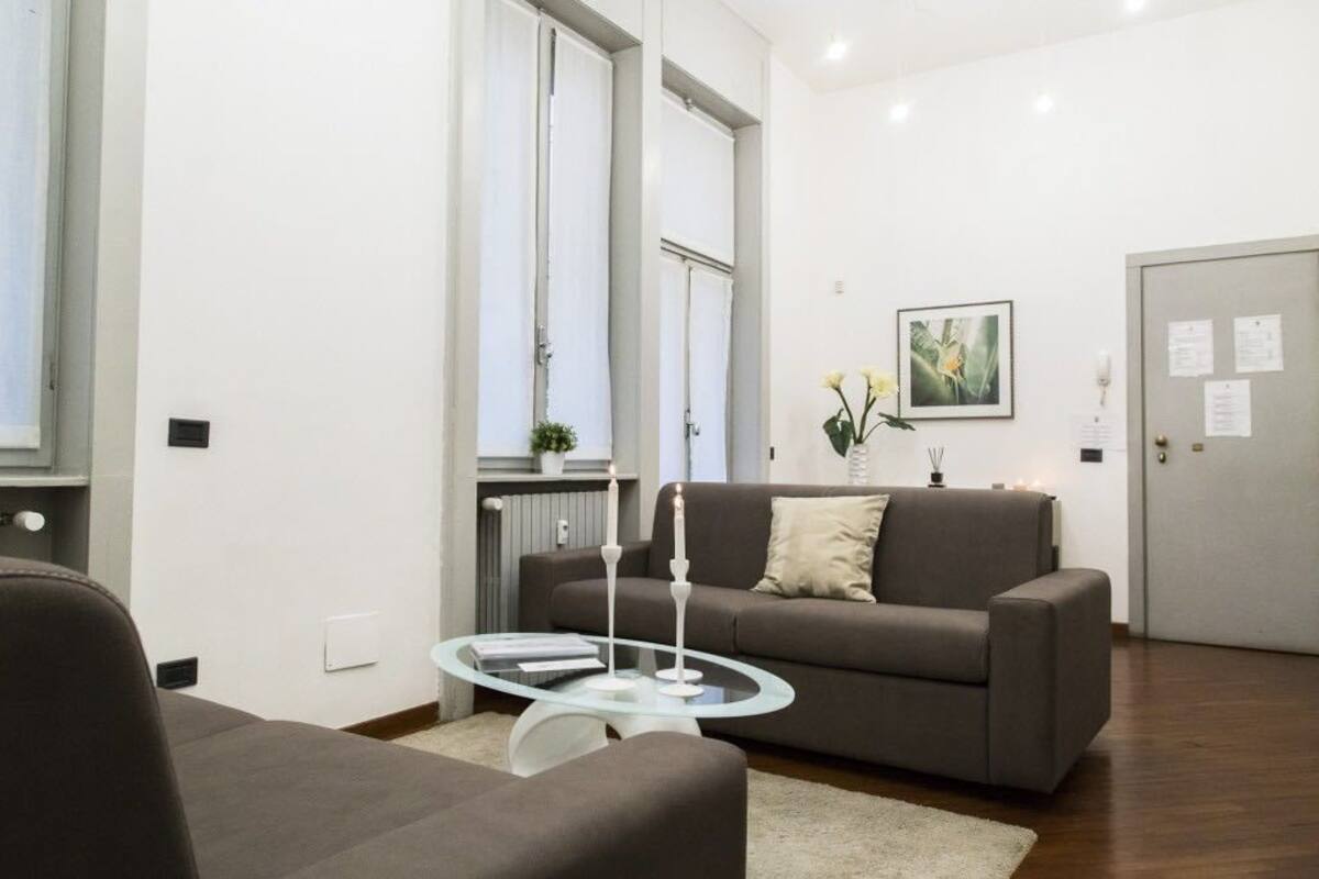 Una sala de estar moderna con un sofá gris, una mesa de centro de vidrio y un sillón blanco. Hay un cuadro en la pared y una puerta con un aviso.