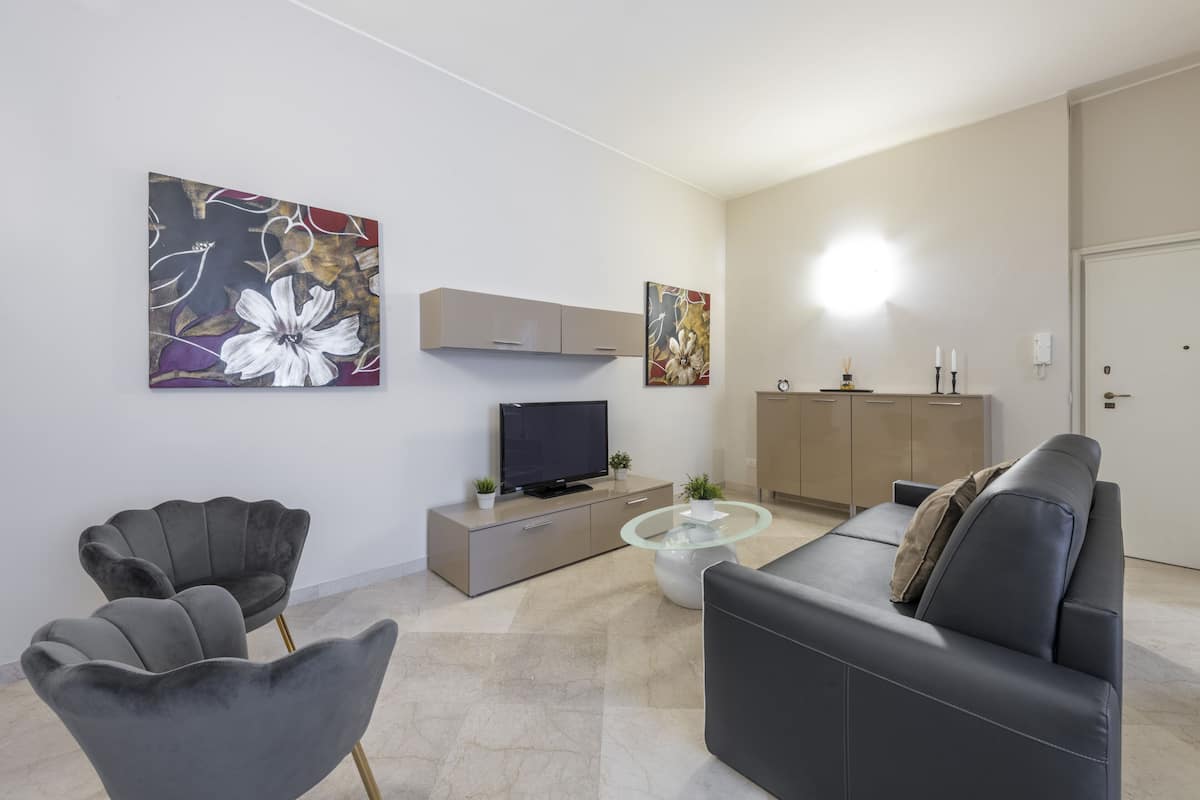 Una sala de estar moderna con un sofá, dos sillones, una mesa de centro de vidrio, un televisor sobre un mueble bajo y obras de arte en las paredes.