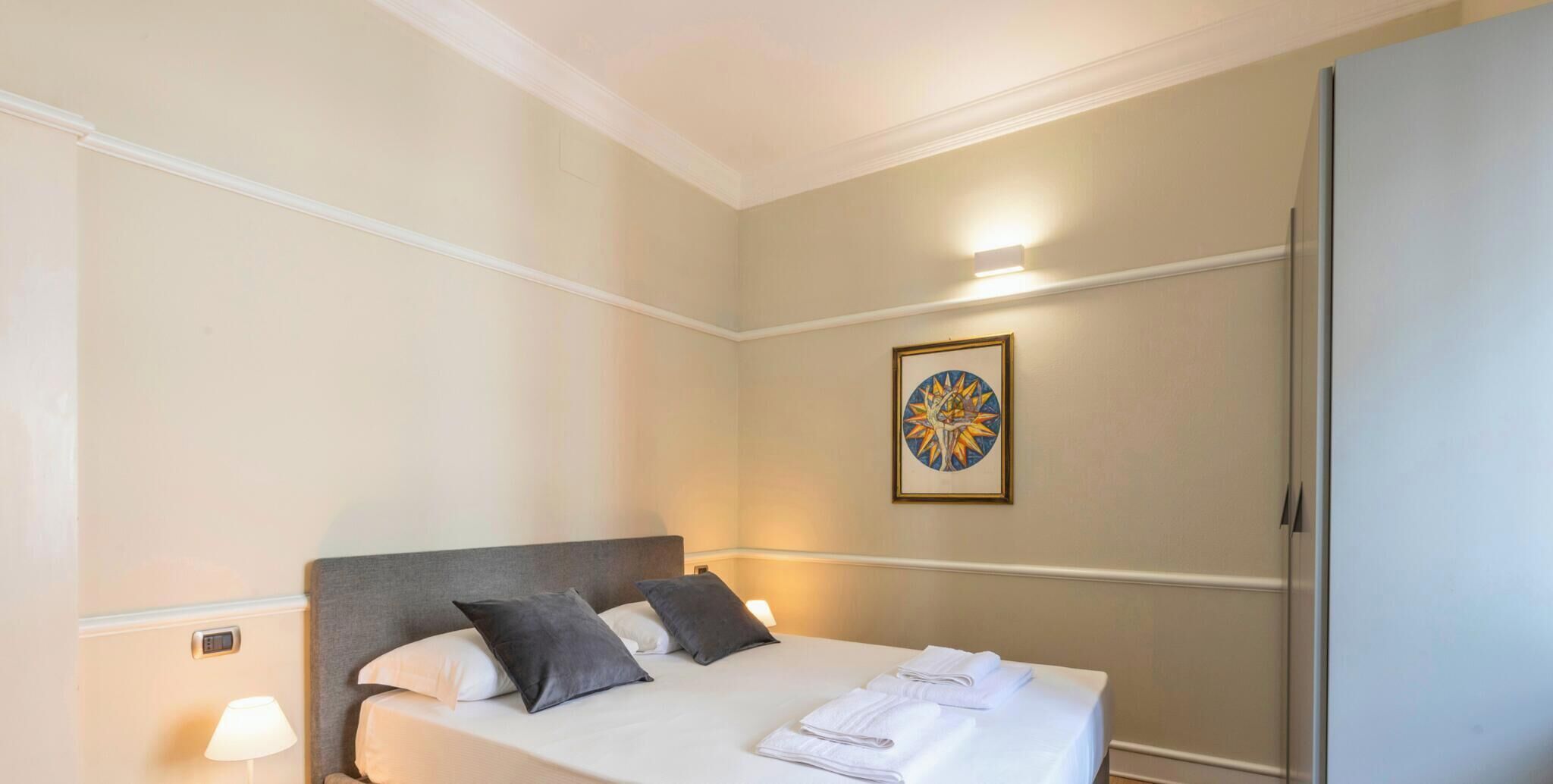Una camera d'albergo con un letto, cuscini, un comodino con una lampada, un'opera d'arte appesa al muro e un armadio.