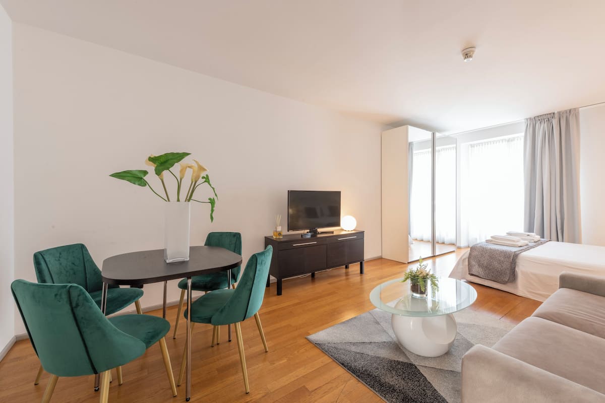 Habitación de hotel moderna con zona de comedor, televisor, cama y un ventanal grande.