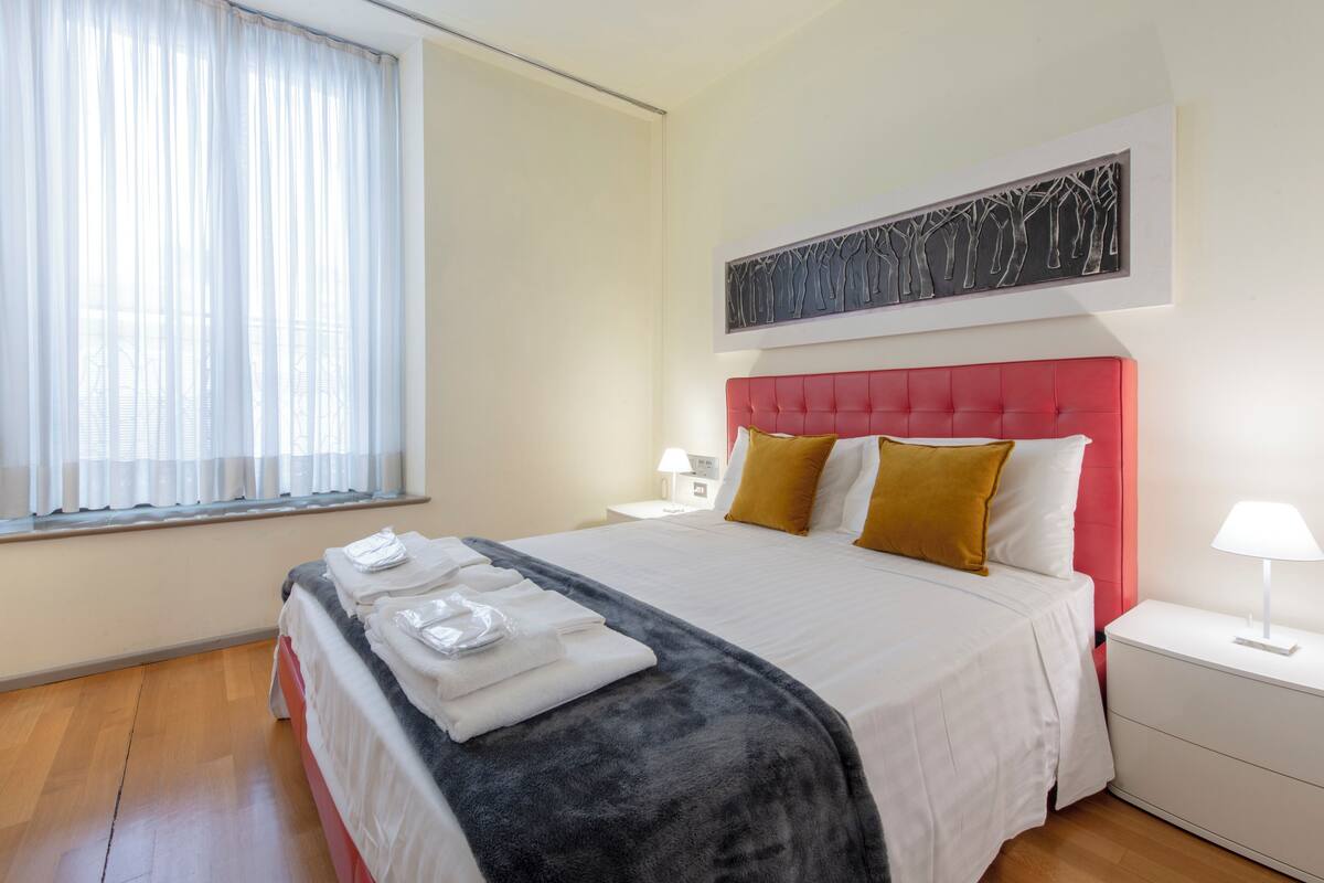 Una cama bien hecha con cabecera roja, ropa de cama blanca y una manta gris. Se ve una ventana con cortinas traslúcidas, una obra de arte en la pared y una mesita de noche con una lámpara.