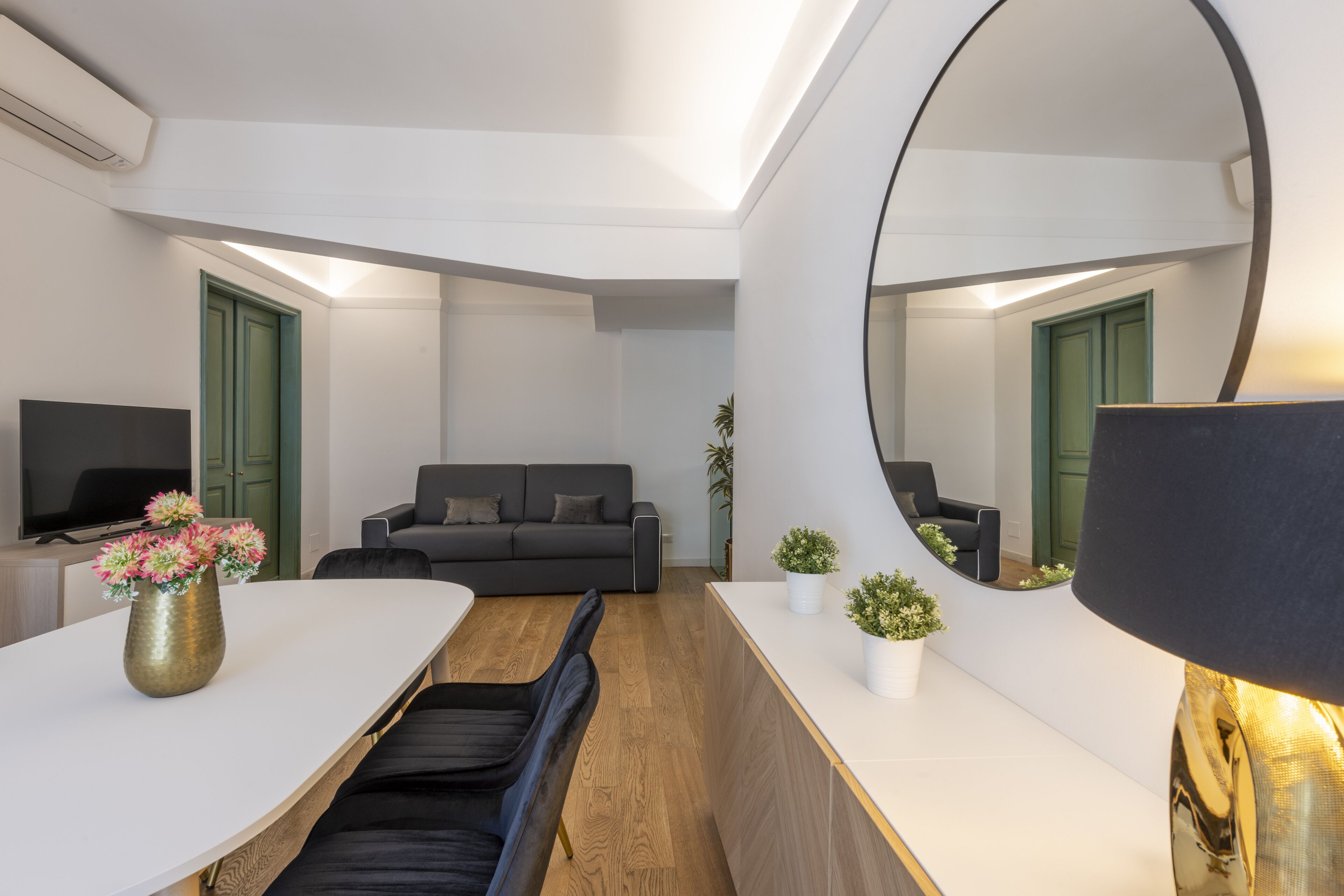 A modern living room with a sofa, a dining table with a vase of flowers, and a large mirror.