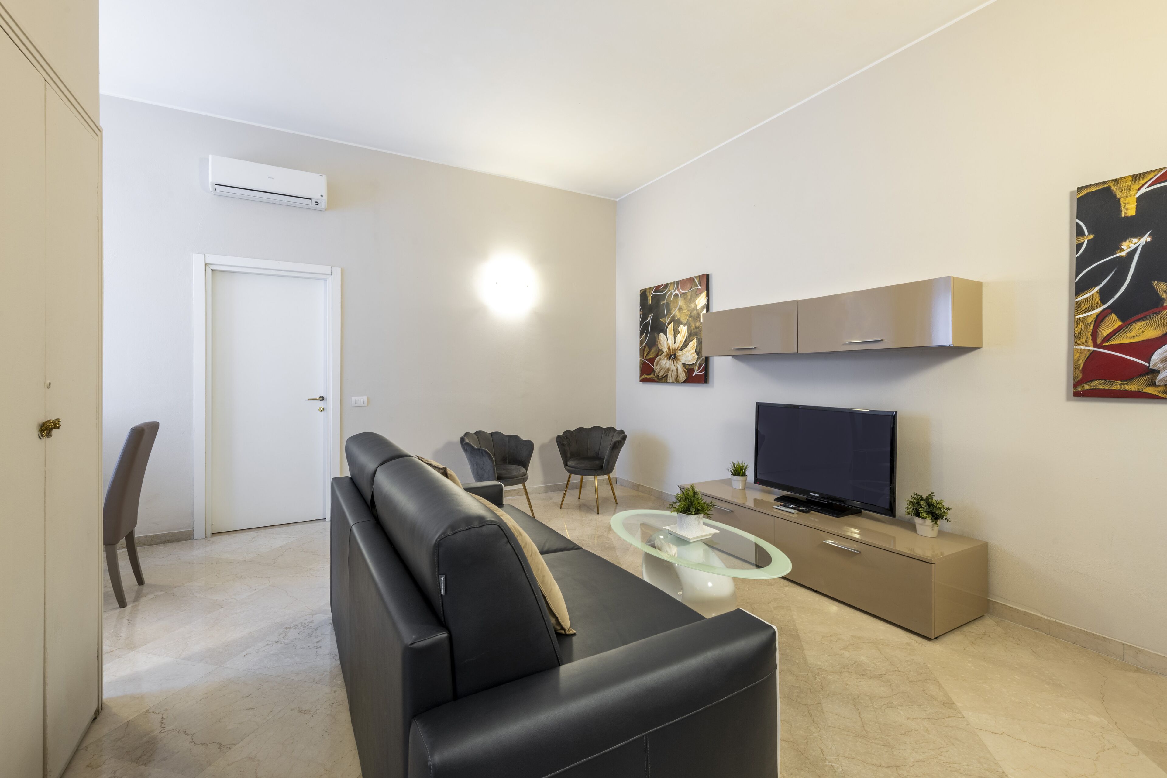 Un salon moderne avec un canapé noir, une table basse en verre, un téléviseur et un meuble. On aperçoit une porte et une chaise au fond.