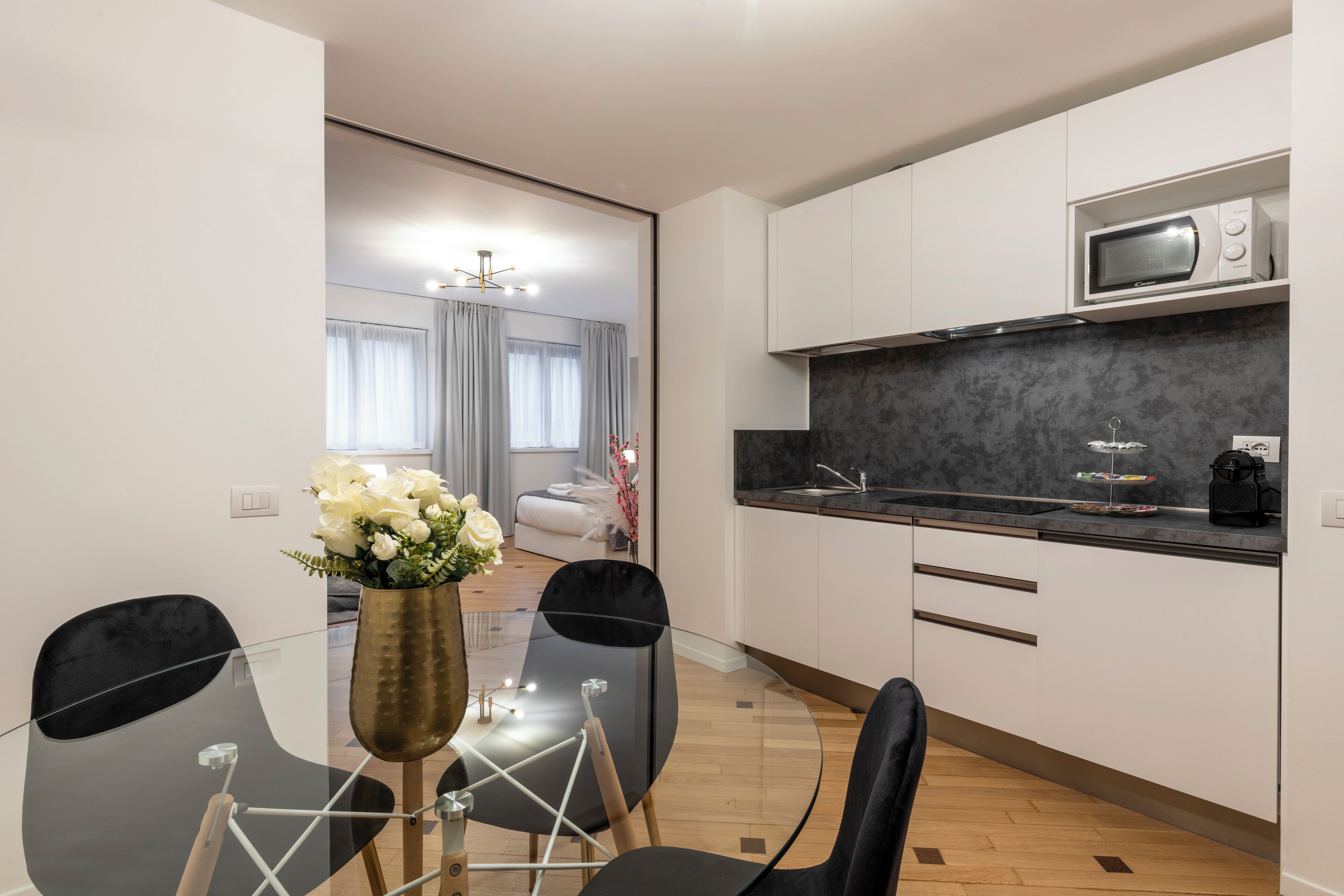 A modern kitchen with white cabinets, a microwave, and a black countertop. There is a dining area with a glass table and black chairs.