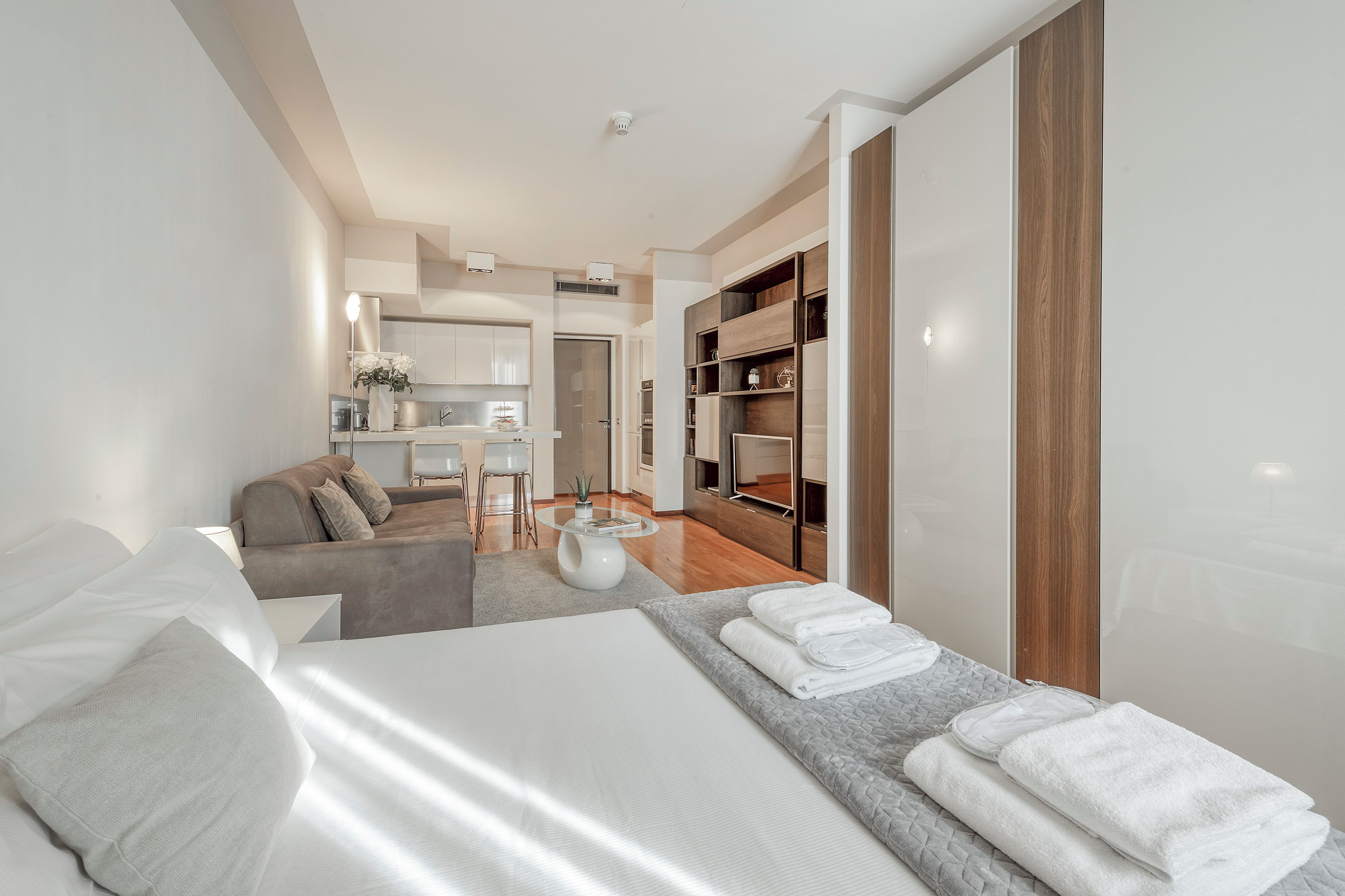 Quarto de hotel moderno com cama, sofá, área de refeições e cozinha ao fundo.
