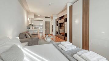 Quarto de hotel moderno com cama, sofá, área de refeições e cozinha ao fundo.