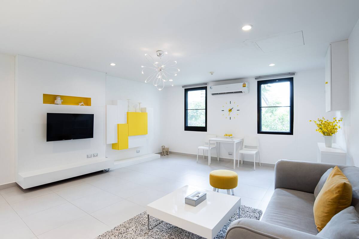 A modern living room with a flat-screen TV, a white coffee table, a sofa with a mustard-colored cushion, and a dining area with a small table and chairs.