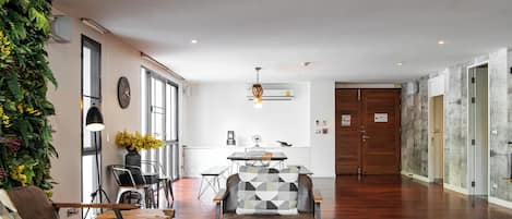 A spacious living area with a wooden coffee table, a sofa, and a dining table. There is a staircase leading to an upper level, and a large window providing natural light.