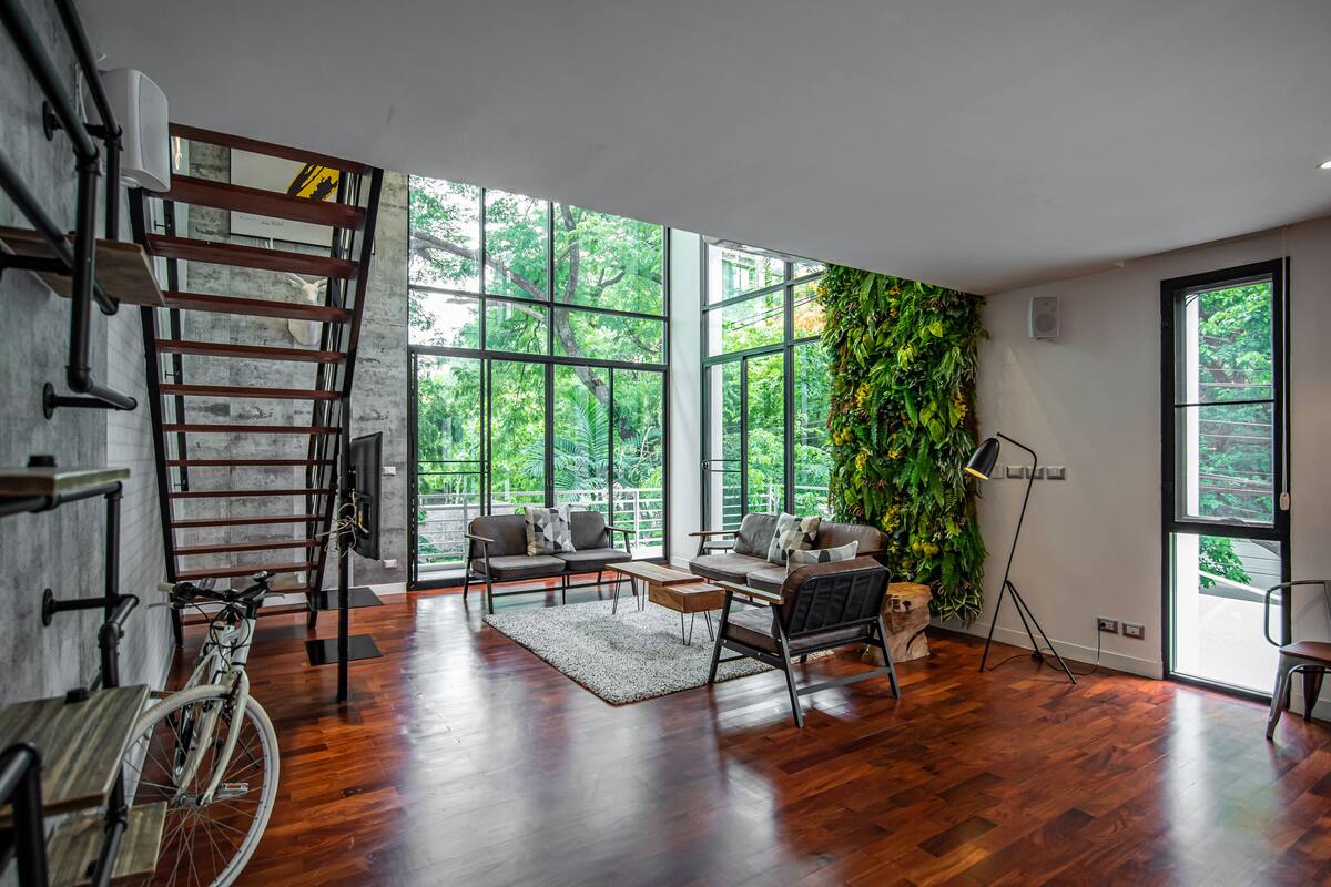 A spacious living room with a wooden floor, large windows, a staircase, and a green wall.
