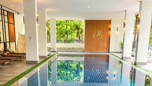 A modern indoor swimming pool with a blue tiled floor, a wooden wall with a logo, and a view of greenery outside. - Benviar Tonson Residence (Bangkok)