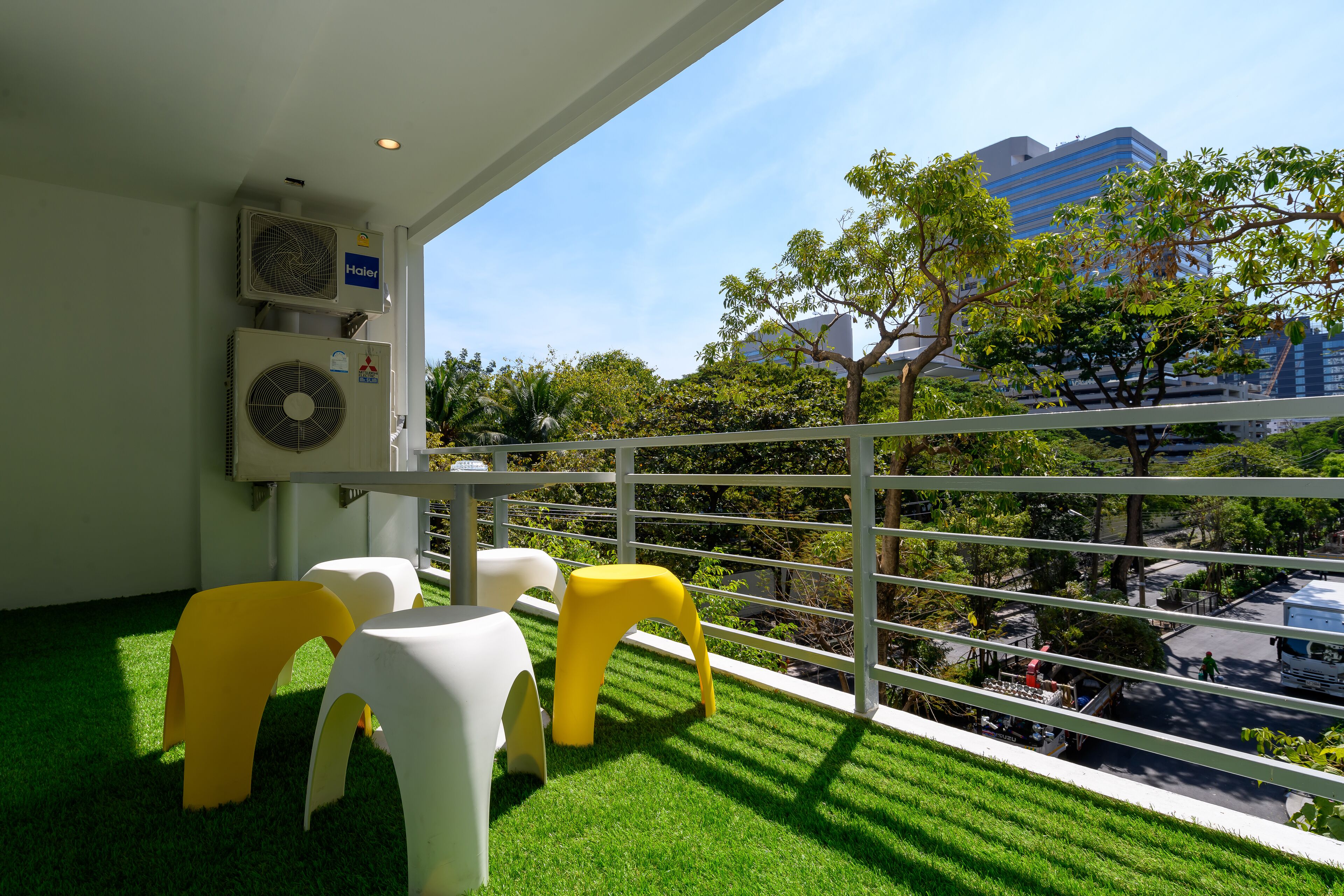 A balcony with artificial grass, white and yellow chairs, and air conditioning units.