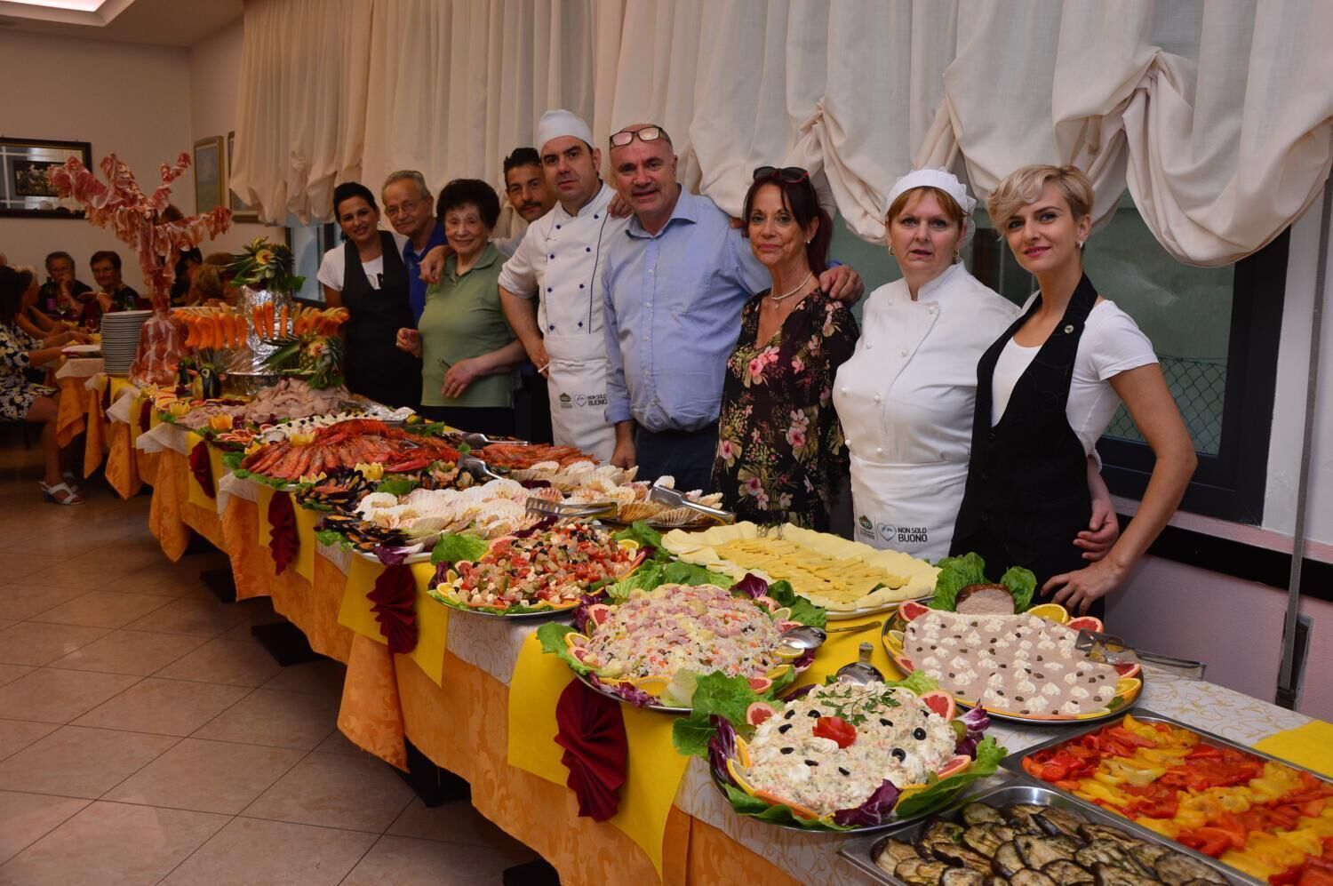 A buffet table with various dishes and a group of people standing behind it.
