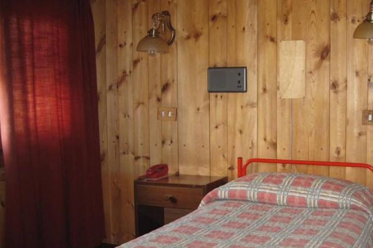 A wooden-paneled room with a bed, a desk, a lamp, and a thermostat.