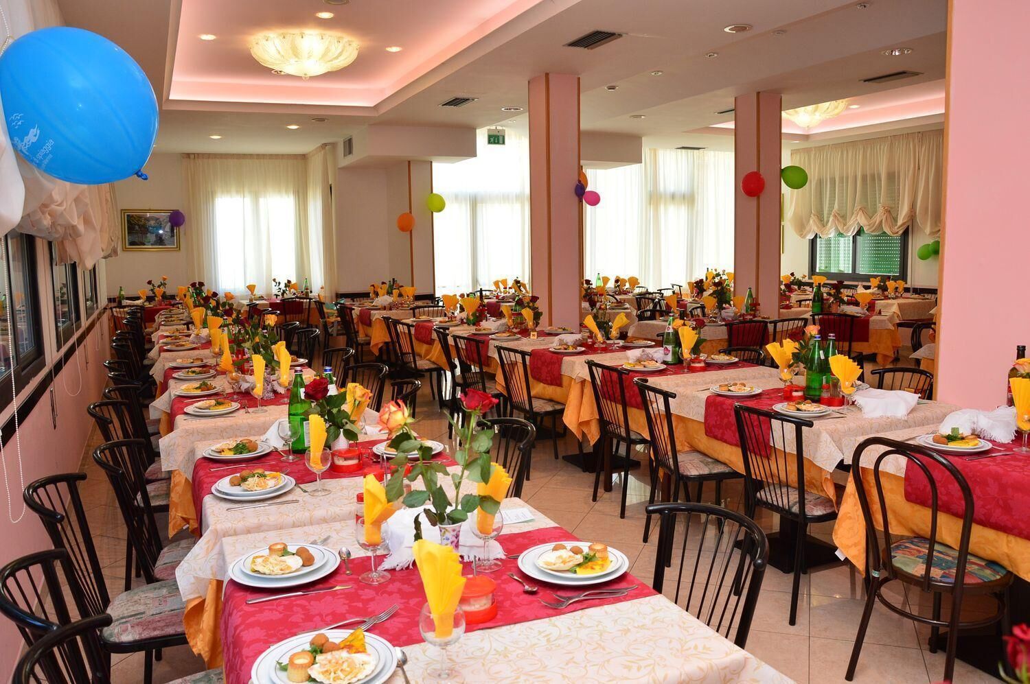 Ampia sala da pranzo con diversi tavoli apparecchiati, tovaglie rosse e gialle, sedie e composizioni floreali al centro.