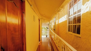 A long, narrow hallway with wooden doors and windows on one side, leading to an open area with a view of greenery.