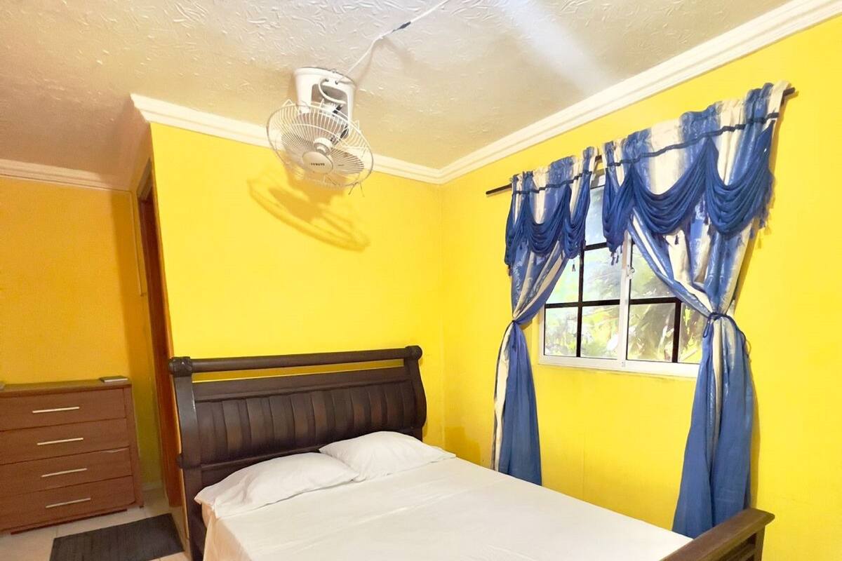 Ein Schlafzimmer mit einem Bett, einem Nachttisch, einer Kommode, einem Fenster mit blauen Vorhängen und einem Deckenventilator.