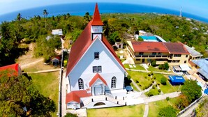 An aerial view of a church with a red roof, surrounded by greenery and a clear view of the ocean.