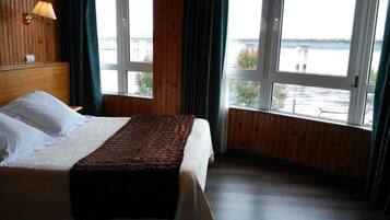 A hotel room with a bed, wooden walls, a window with a view of a body of water, and a lamp on the wall.