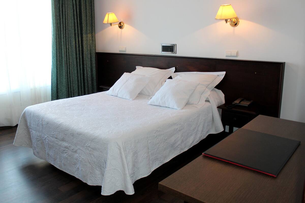 Cama bem arrumada com colcha e travesseiros brancos, mesa de cabeceira com telefone e um caderno vermelho, e duas luminárias fixadas na parede.
