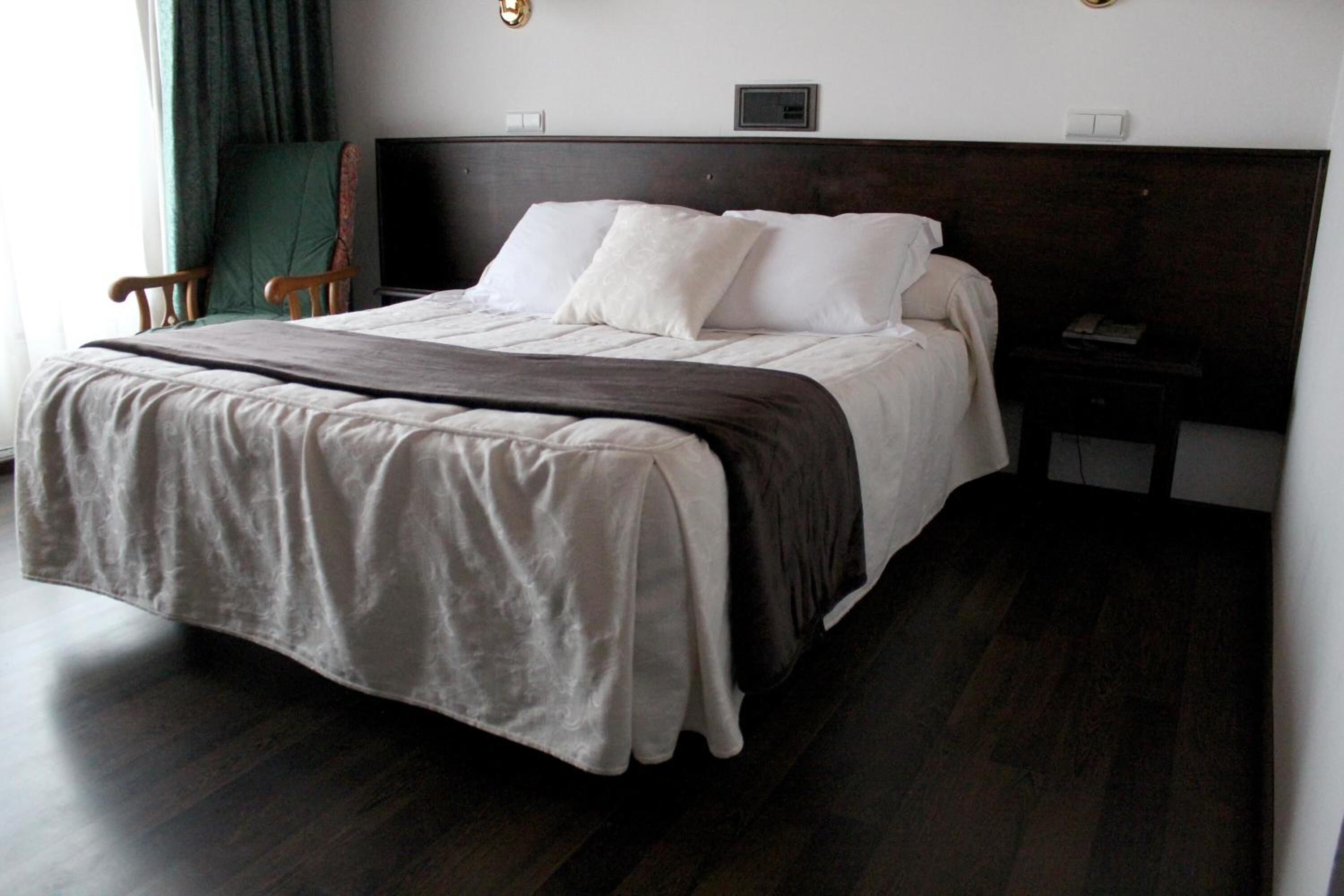 Una cama bien tendida con un cabecero oscuro, una mesita de noche con una lámpara y una silla junto a la ventana.